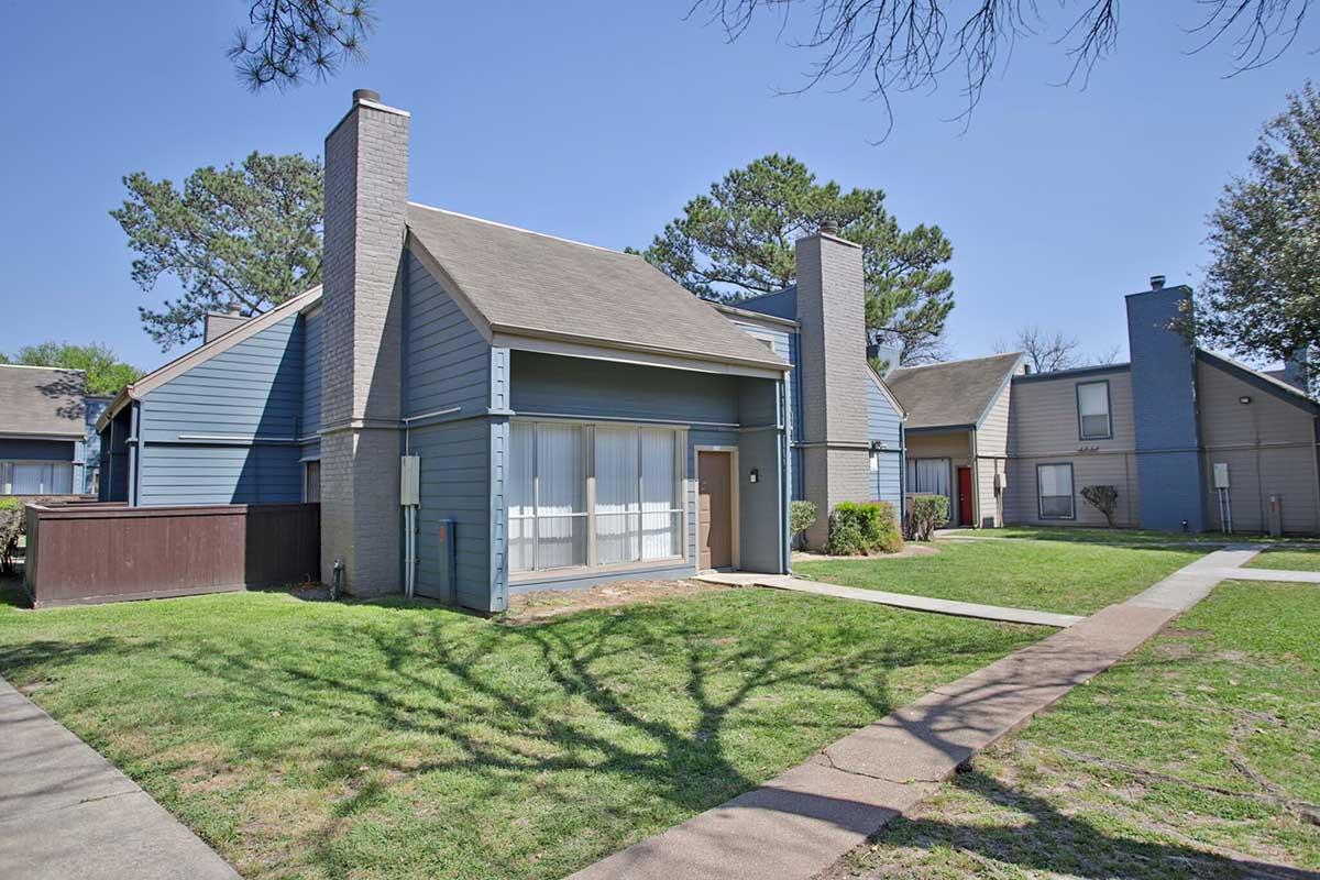 Exterior view of a two-story townhouse complex. The buildings have blue siding, asphalt roofs, and large windows. Lush green grass and trees surround the area, with walkways leading to the entrances of the homes. Clear blue sky in the background.