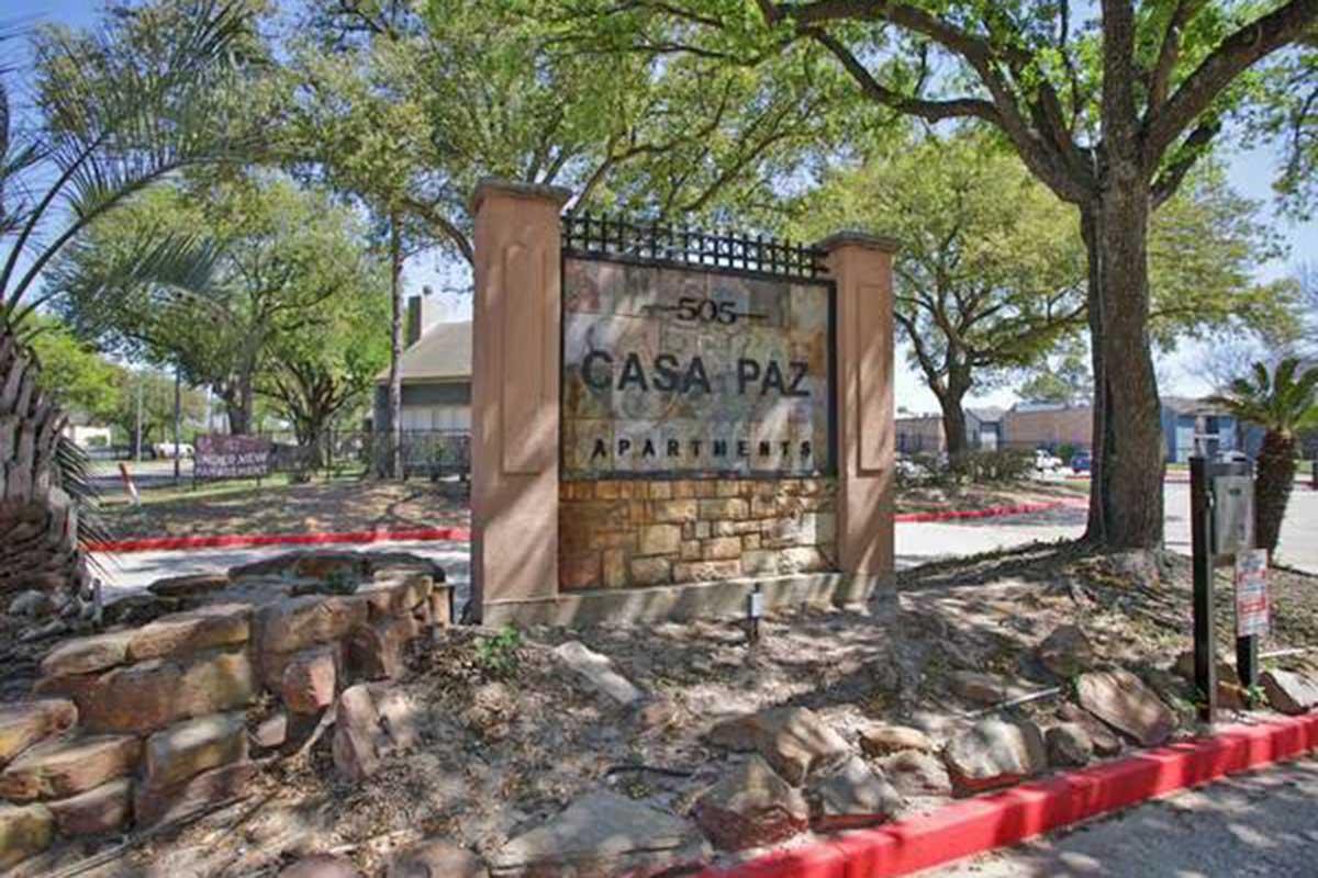 Sign for Casa Paz Apartments, featuring a stone base and prominently displaying the name "Casa Paz" along with the address "505." Surrounded by trees and landscaping, the image depicts a welcoming entrance to the apartment complex, with a red curb and nearby signage visible.