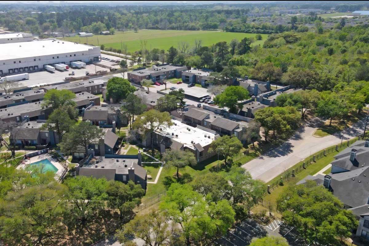 Aerial view of a suburban residential complex surrounded by greenery. The layout includes several multi-unit buildings, a pool area, and a parking lot. Open fields are visible in the background.