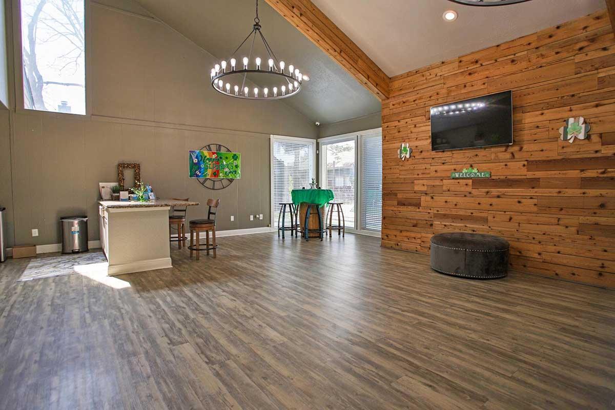Bright and spacious interior of a modern room featuring a wooden accent wall, large windows, and a high ceiling. A stylish chandelier hangs above a table with chairs, while a TV is mounted on the wall. A green table is visible in the background, along with a cozy ottoman.