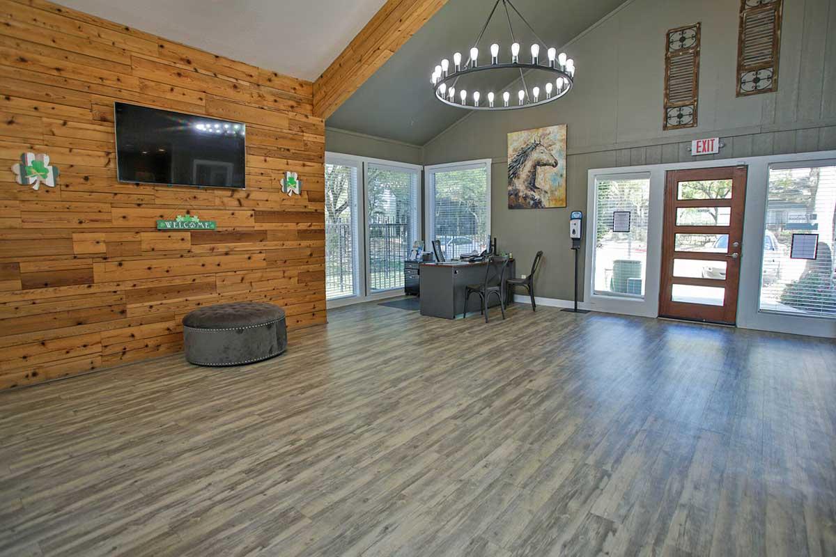 Interior of a welcoming reception area with wooden walls, a large circular chandelier, a flat-screen TV on the wall, and a desk with a chair. Large windows provide natural light. A horse-themed painting is visible on the wall, and there is a round seating area with a cushion. Entrance doors are visible.