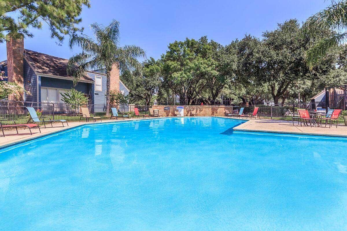 A sunny pool area featuring a bright blue swimming pool surrounded by green trees and shrubs. Colorful lounge chairs are arranged around the pool, with a residential building visible in the background. The clear sky reflects a warm atmosphere, inviting relaxation and leisure.