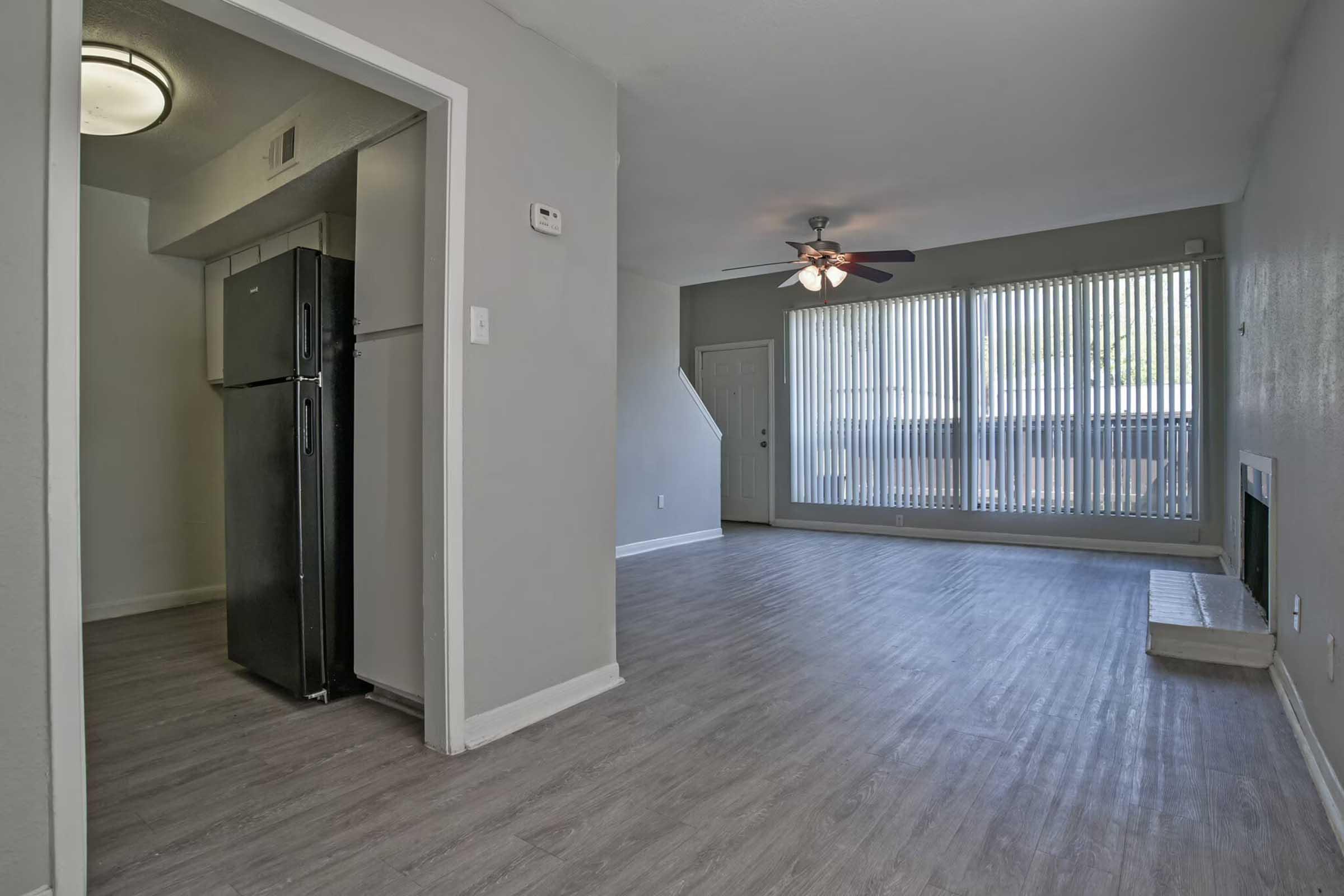 Interior view of a modern living space featuring a light gray color scheme, hardwood-style flooring, and large windows with vertical blinds. In the background, there is a kitchen with a black refrigerator, and a door leading outside. The room is well-lit and spacious, creating an inviting atmosphere.