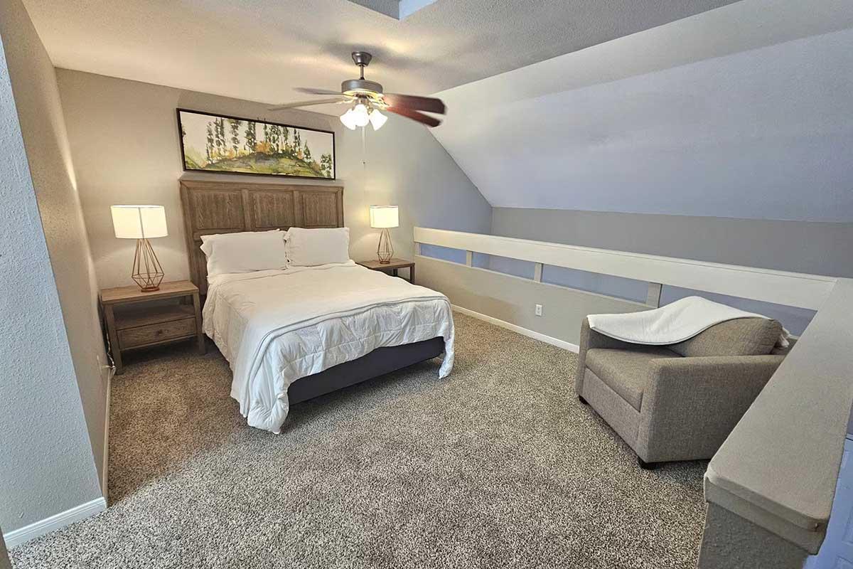 A cozy bedroom featuring a queen-sized bed with white bedding, a wooden headboard, and two bedside lamps. The room has a light-colored wall and carpet, a ceiling fan, and a plush armchair in the corner. A painting of trees hangs above the bed, adding a touch of decor to the serene atmosphere.