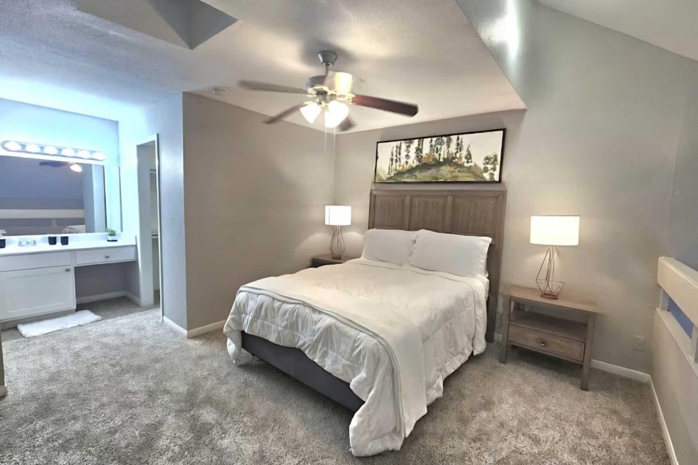 A cozy bedroom featuring a queen-sized bed with a white comforter, two bedside lamps, and a wooden nightstand. A ceiling fan hangs above, and a large piece of artwork is displayed on the wall. A lighted vanity area is visible in the background, with a mirror and additional storage. The room has soft neutral tones and carpeted flooring.