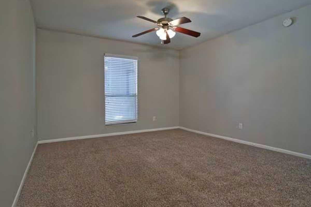 A neutral-colored room with a ceiling fan and carpeted floor. A window with blinds allows natural light to enter, illuminating the spacious area. The walls are plain, enhancing the room's simplicity and versatility for various uses.