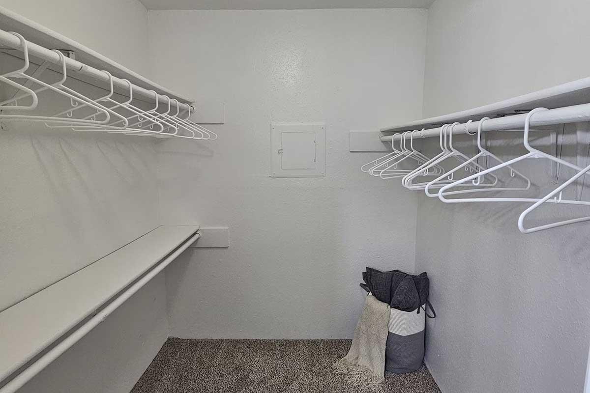 A clean, empty closet with two rows of white wire hangers on either side and a shelf for additional storage. The floor is carpeted, and a folded blanket is placed against the wall in a gray and white basket.