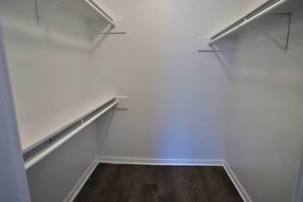 Empty closet with plain white walls, featuring two hanging rods along the sides and a shelf along the back wall. The floor is dark, adding contrast to the light-colored walls. There are no items or personal belongings visible inside.