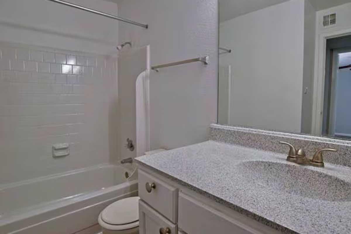 A clean bathroom featuring a white tiled bathtub and shower, a light-colored countertop with a sink, and a wall with a towel bar. The space is illuminated and has a simple, modern design. There is also a door leading to another room visible in the background.
