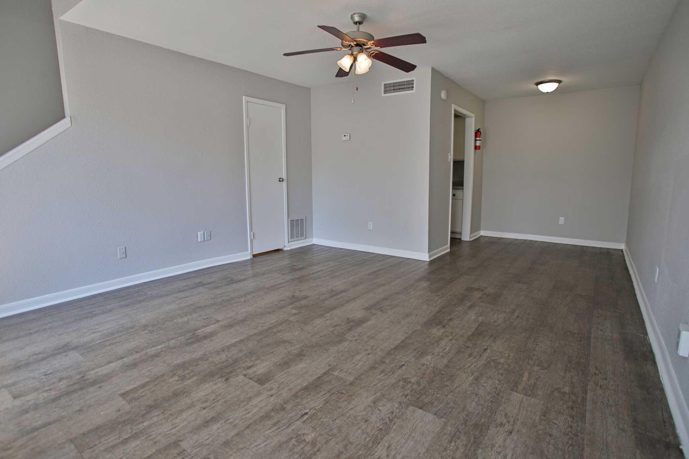 Empty living room with light gray walls and a wooden floor. A ceiling fan hangs in the center, and there is a door on the left leading to another room. The space is well-lit with natural light and has a clean, modern feel. The kitchen is partially visible through an open doorway.