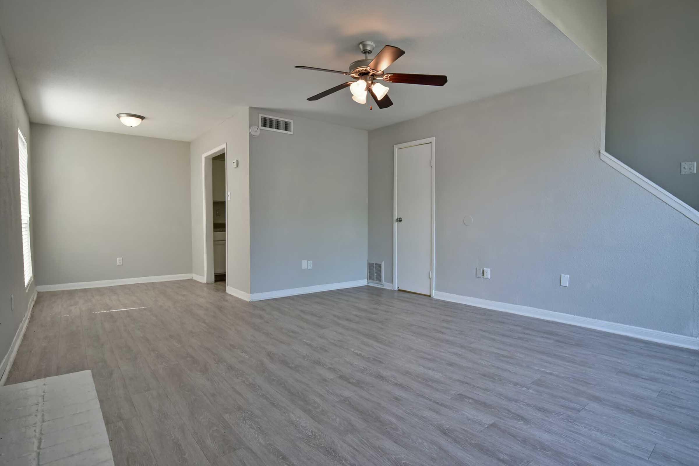 Spacious, empty living room with light gray walls and wood-like flooring. A ceiling fan with three blades hangs from the ceiling. Natural light enters through a window, and there is a doorway leading to another room. A staircase is visible in the background, adding depth to the space.