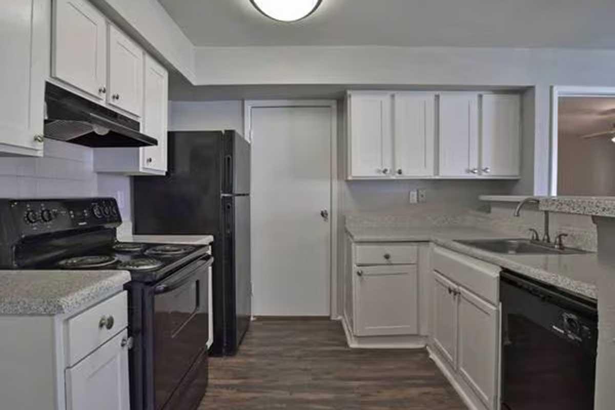 Modern kitchen featuring white cabinetry, granite countertops, a black refrigerator, a black oven with a stovetop, and a sink. The space has a light-colored wall, wooden flooring, and natural light from an overhead fixture. A door leads to another area, enhancing the open layout.