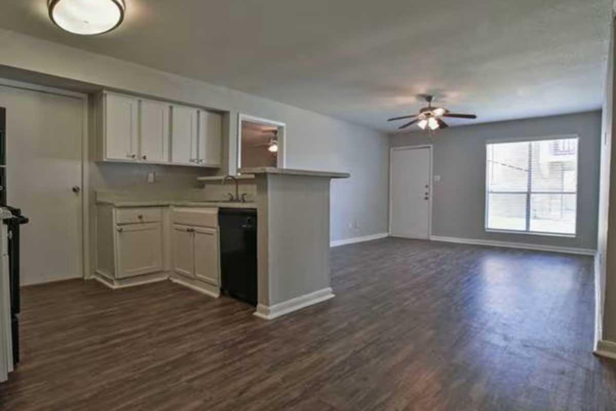 Interior view of a modern apartment featuring an open floor plan. The living area includes a ceiling fan and large windows providing natural light. A kitchen area with white cabinets and black appliances is visible, alongside a light-colored wall and laminate flooring.