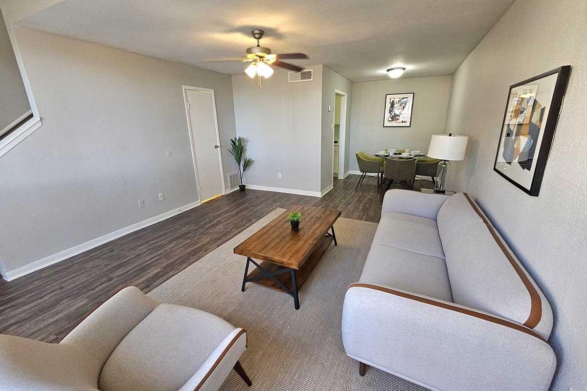 A bright and spacious living room featuring a light gray sofa with wooden legs, a wooden coffee table with a small potted plant, and a ceiling fan. The walls are light-colored, and there is a doorway leading to another room with a dining area visible in the background.