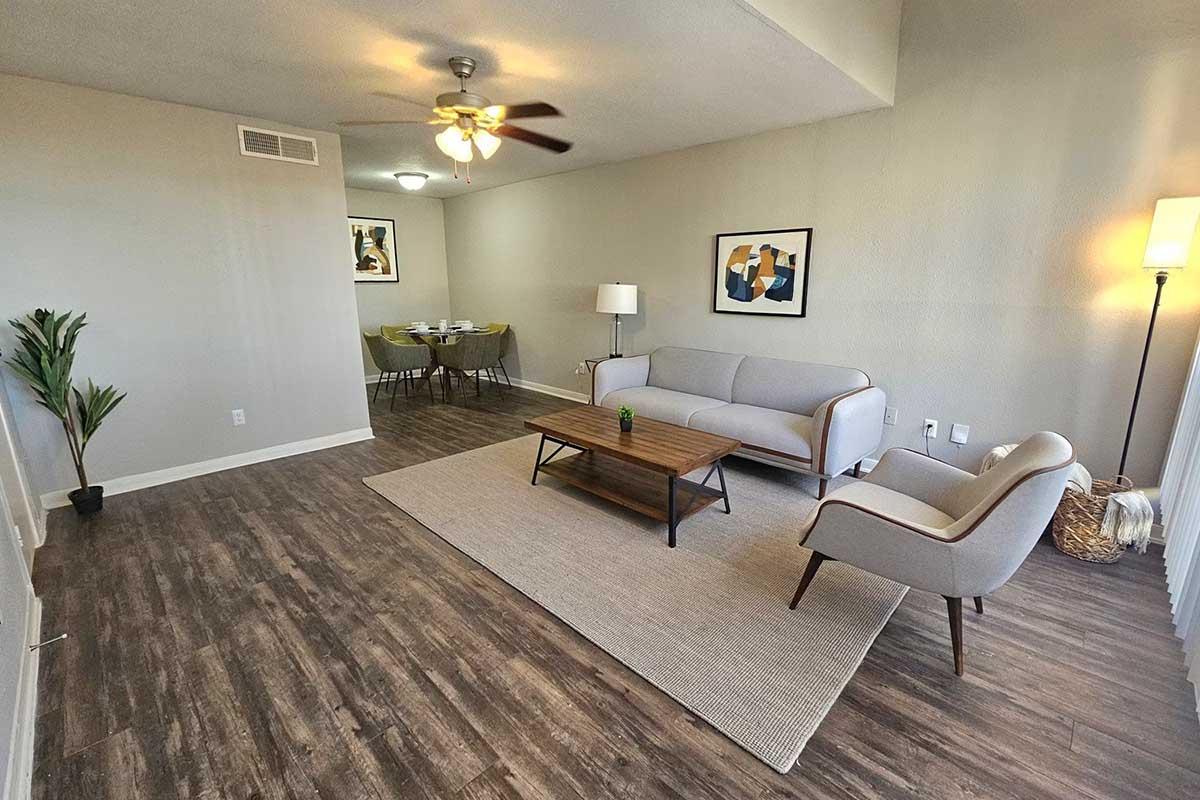A modern living room featuring a light gray sofa and a matching armchair beside a wooden coffee table. The room has a decorative plant, a ceiling fan, and artwork on the walls. A dining area is visible in the background with a table set for four. The flooring is a warm brown color, adding to the cozy atmosphere.