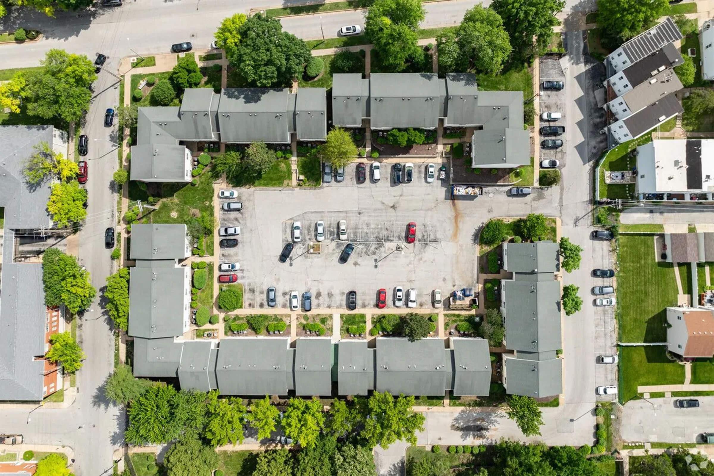 Aerial view of a residential complex with multiple buildings arranged in a square around a central parking area. Surrounding streets are lined with trees and parked cars, showcasing a mix of residential homes and nearby structures. The scene captures greenery and urban development.