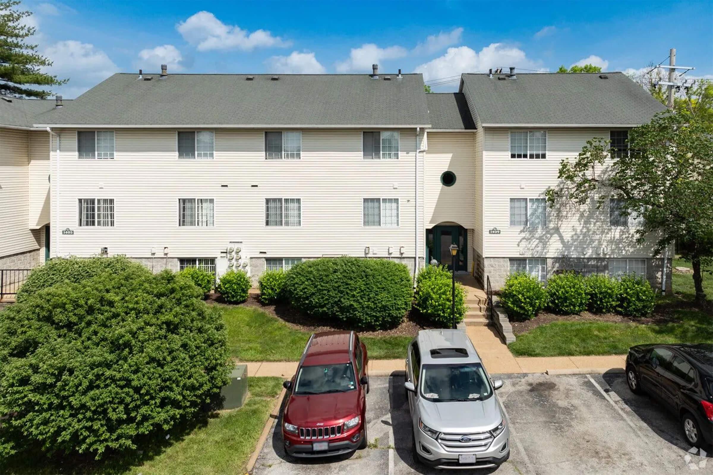 View of a two-story residential building with light-colored siding surrounded by green bushes and trees. In the foreground, there are several parked cars, including red and silver vehicles, stationed on a paved driveway. The sky is blue with some clouds, indicating a sunny day.