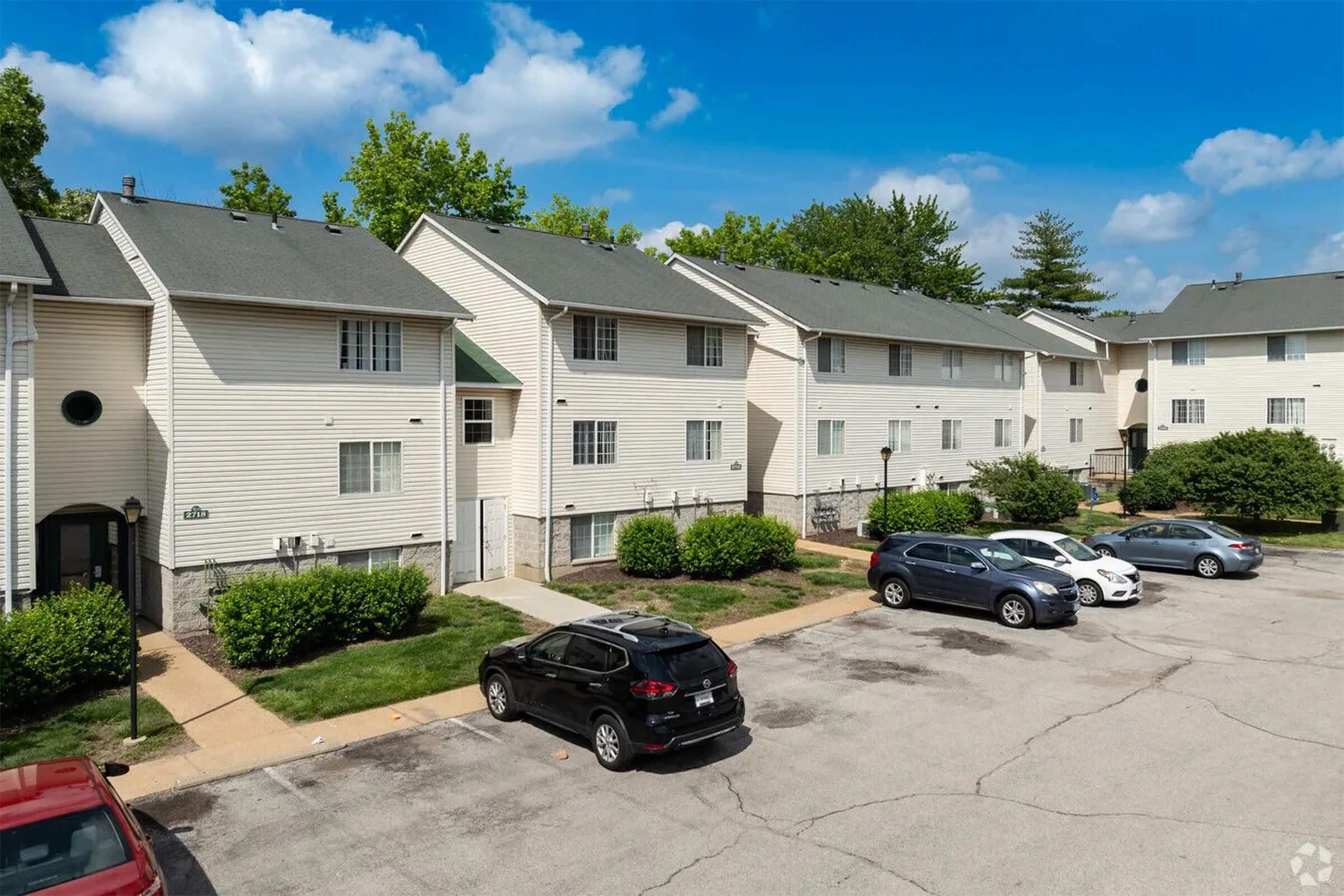 Aerial view of a residential apartment complex featuring beige two-story buildings, well-maintained greenery, and a parking lot with several cars. The sky is clear with scattered clouds, providing a bright and inviting atmosphere.