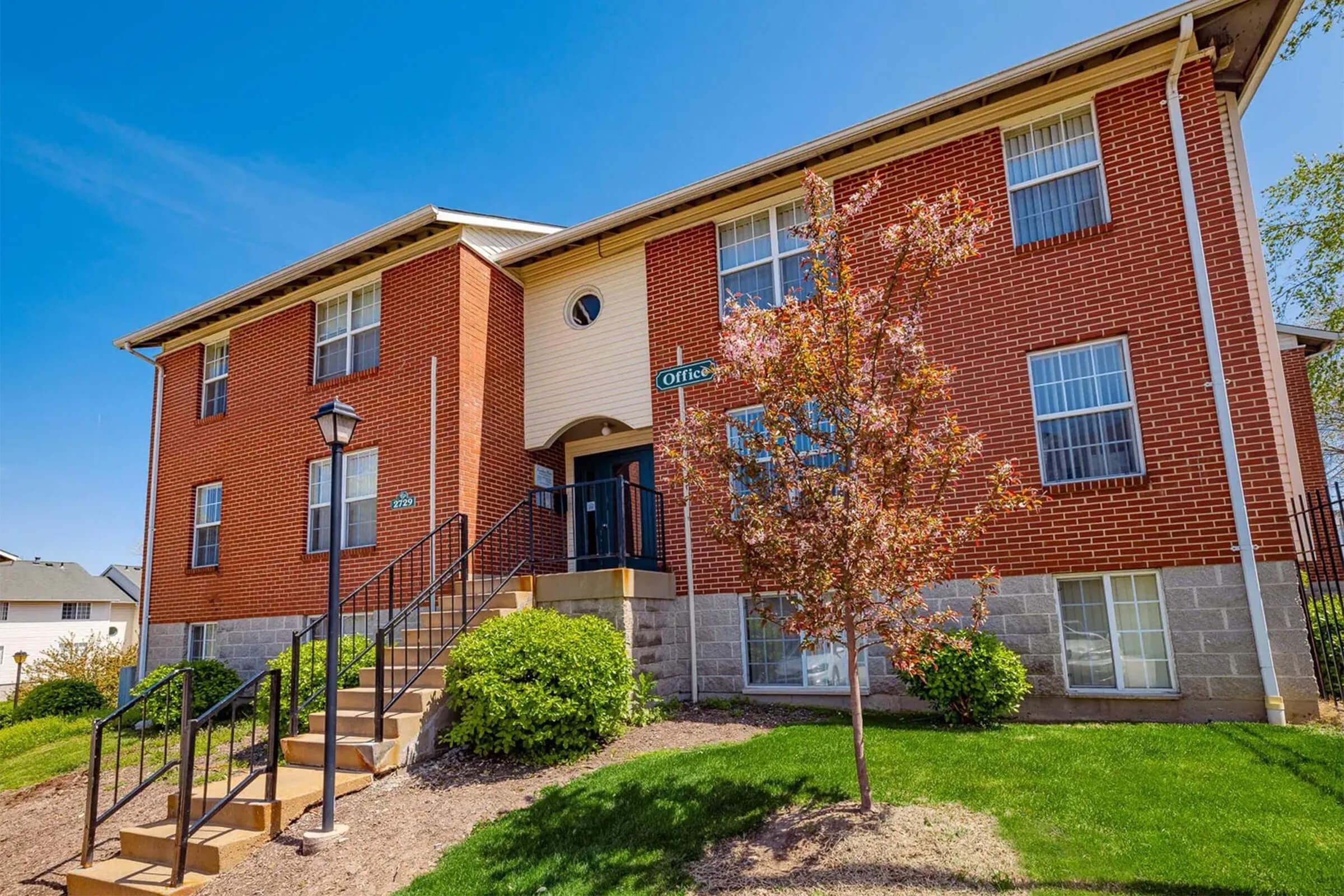 A two-story brick apartment building featuring a staircase leading to the front entrance with a sign that reads "Offices." The building is surrounded by green grass and small shrubs, under a clear blue sky. A decorative lamp post stands in front, adding to the inviting appearance.