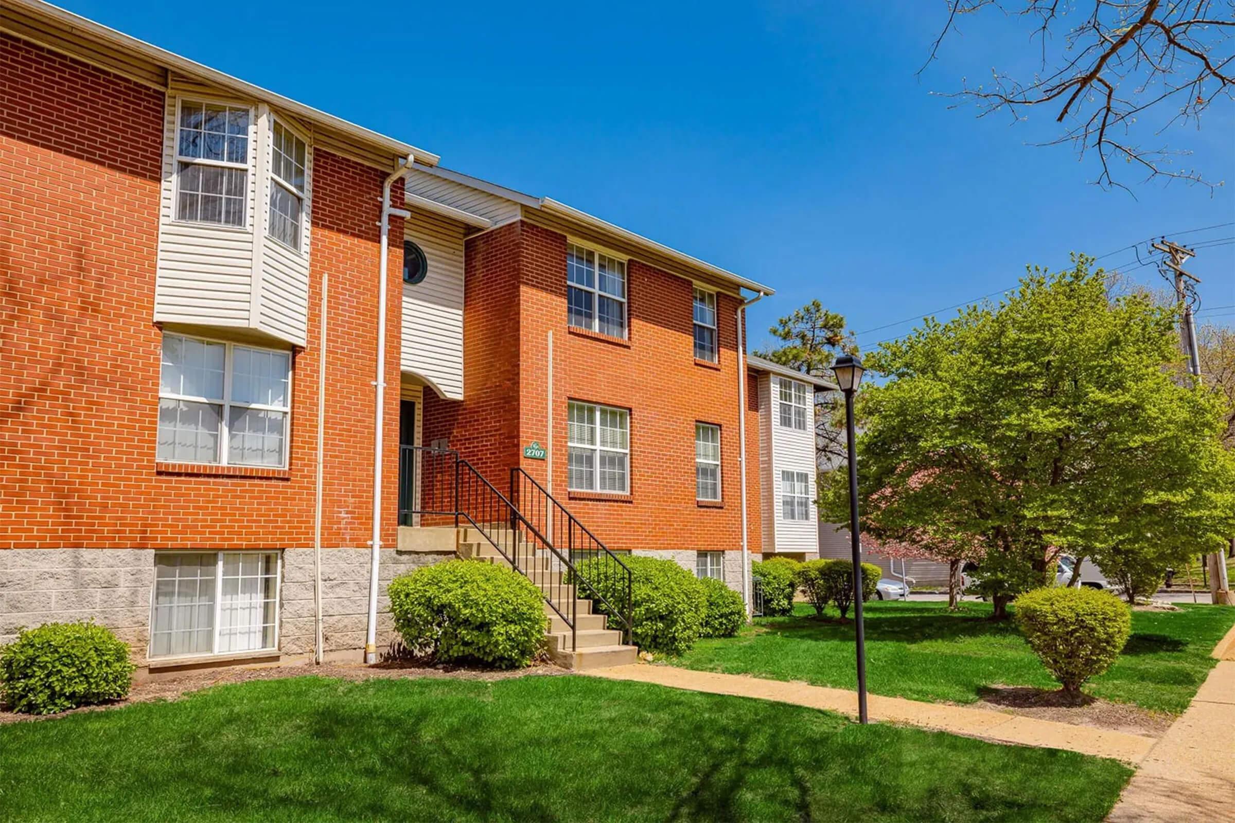 A red brick apartment building with multiple windows and a staircase leading up to the entrance. Lush green grass and neatly trimmed bushes surround the building. The sky is clear and bright blue, indicating a sunny day.