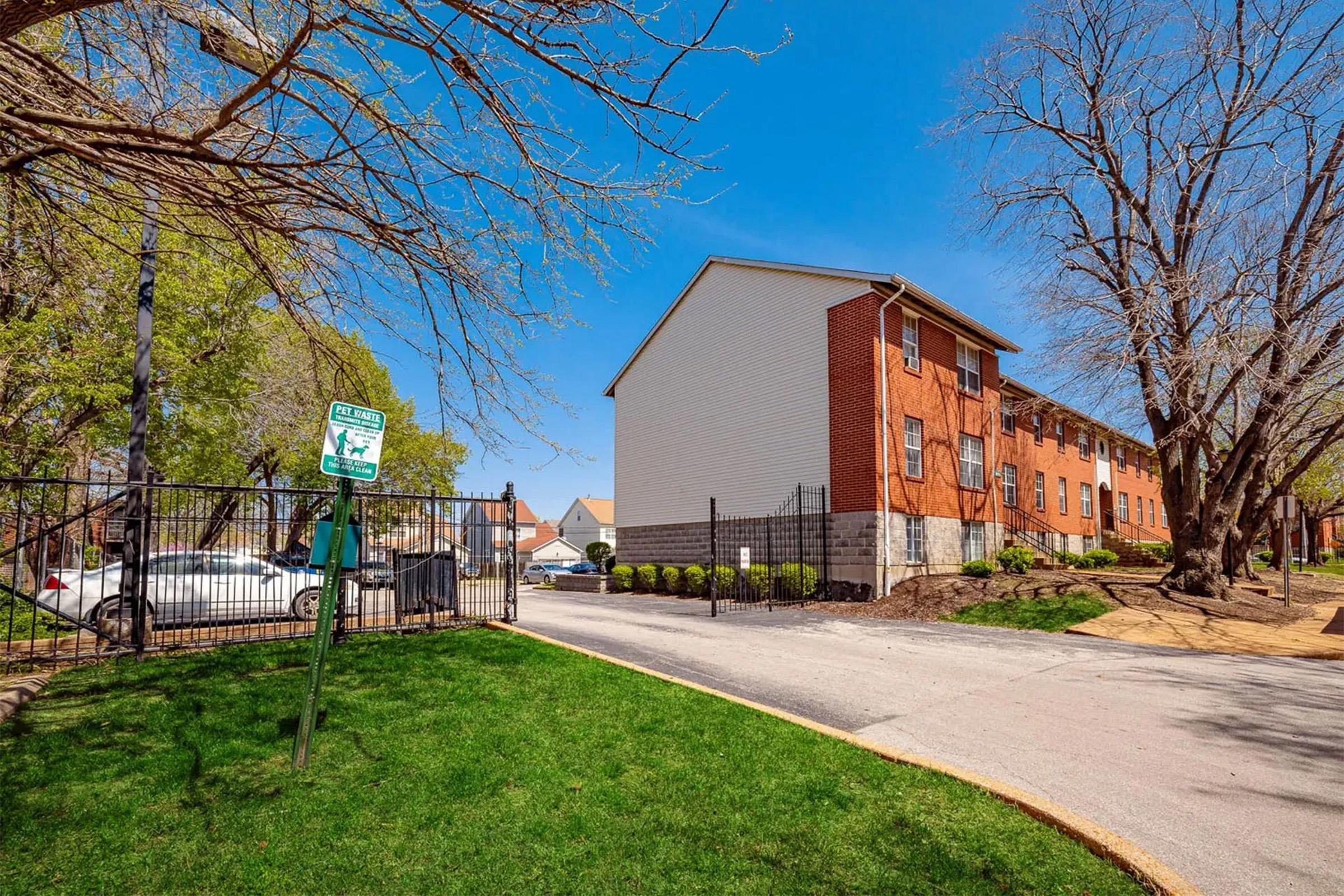 A residential area featuring a brick building surrounded by trees and a green lawn. There is a sign indicating a pet area nearby, and the scene includes a driveway leading to parked cars behind a gated fence under a bright blue sky.