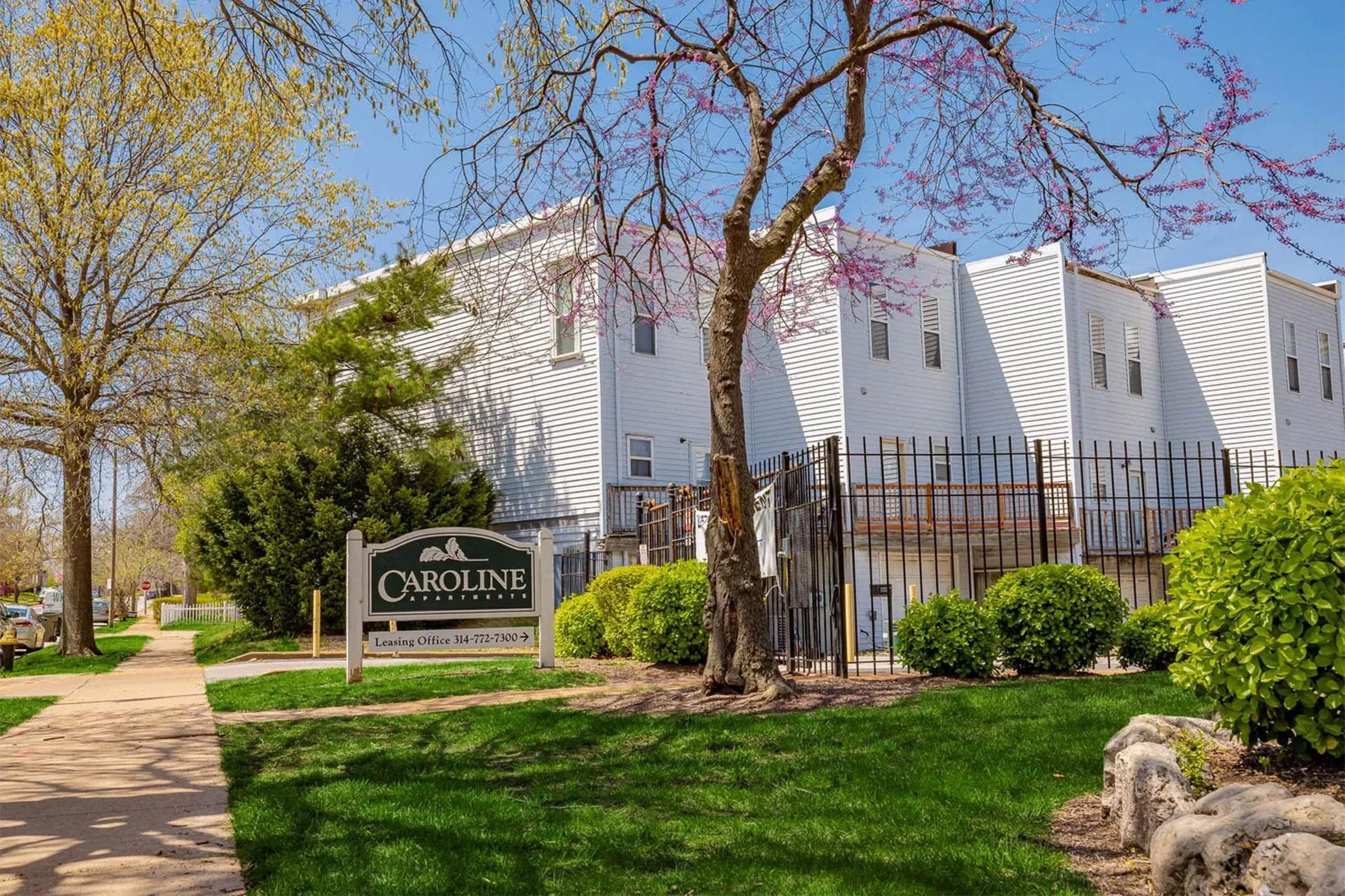 A sunny outdoor view of a residential building with a sign that reads "Caroline" for leasing information. The building features white siding and is surrounded by well-maintained greenery and trees. A clear blue sky enhances the inviting atmosphere of the area.