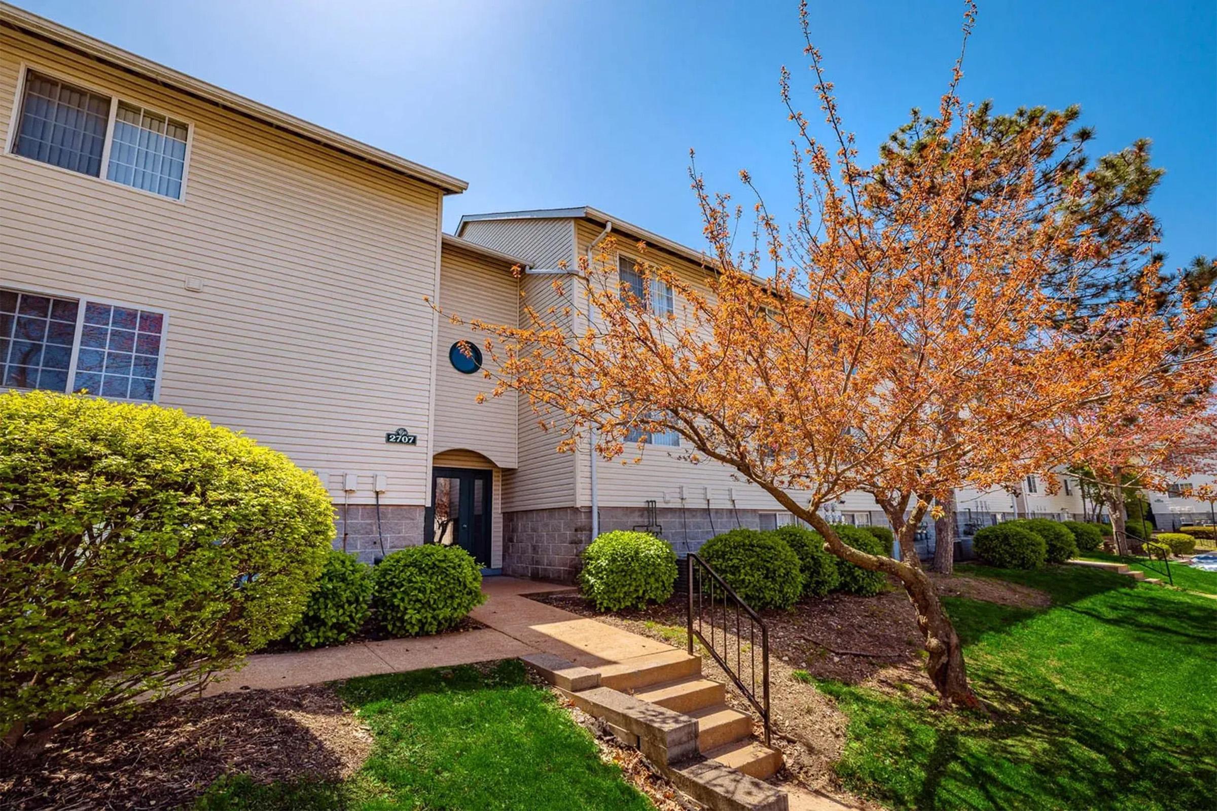 A well-lit image of a two-story apartment building surrounded by green grass and shrubs. A vibrant flowering tree is in the foreground, adding color to the scene. The building has several windows and a front entrance with stairs leading up, under a clear blue sky.