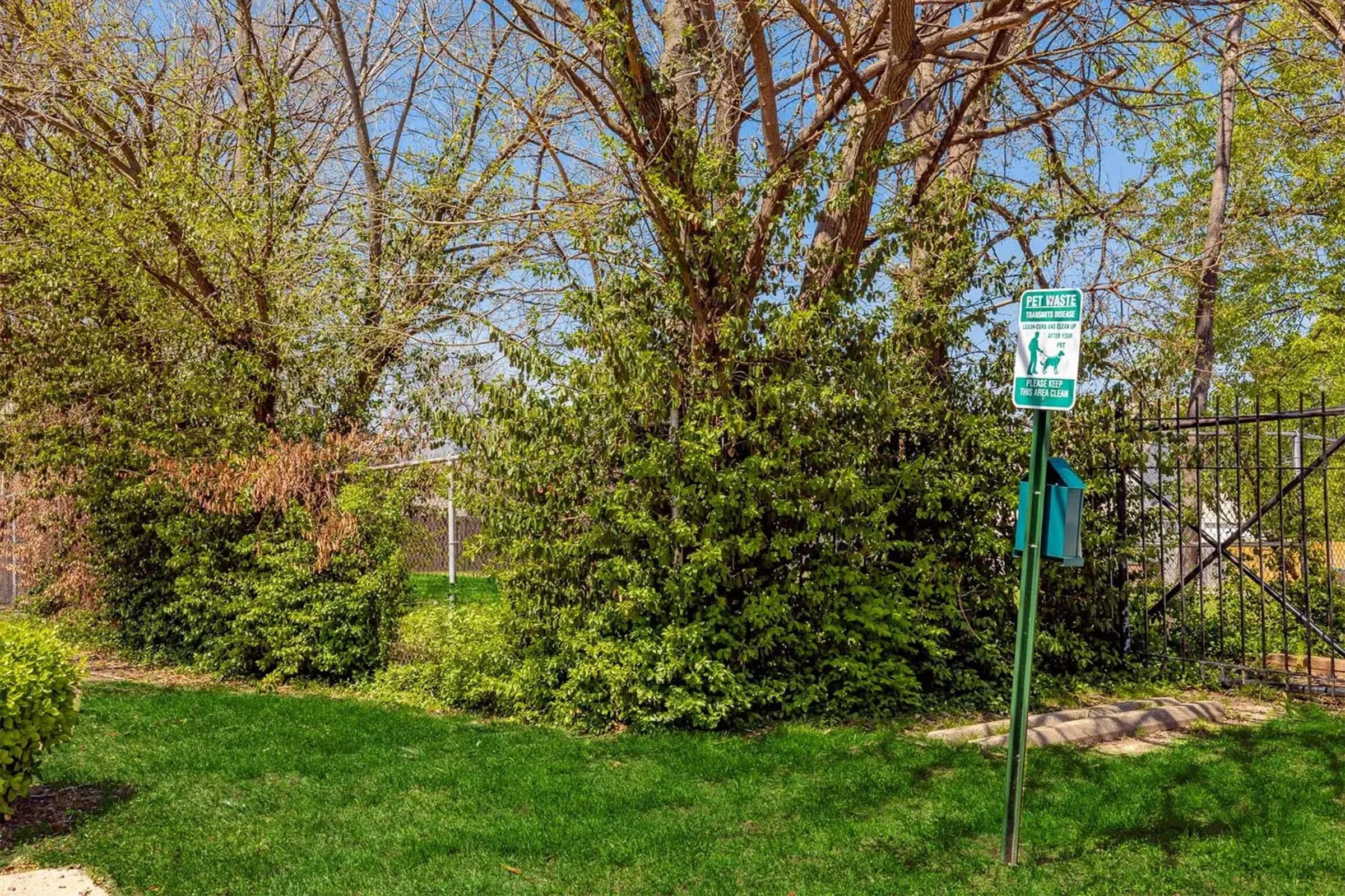 A grassy area with trees and shrubs. A green sign indicating a pet area is visible, along with a fenced section in the background. The sky is clear and blue, suggesting a pleasant day.