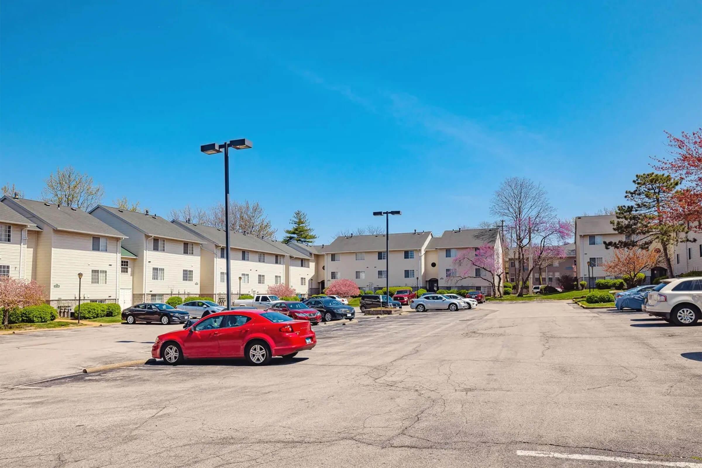 A spacious parking lot filled with cars, including a red sedan, surrounded by several multi-story apartment buildings. The scene is set under a clear blue sky with a few trees and flowering plants nearby, indicating a pleasant day.