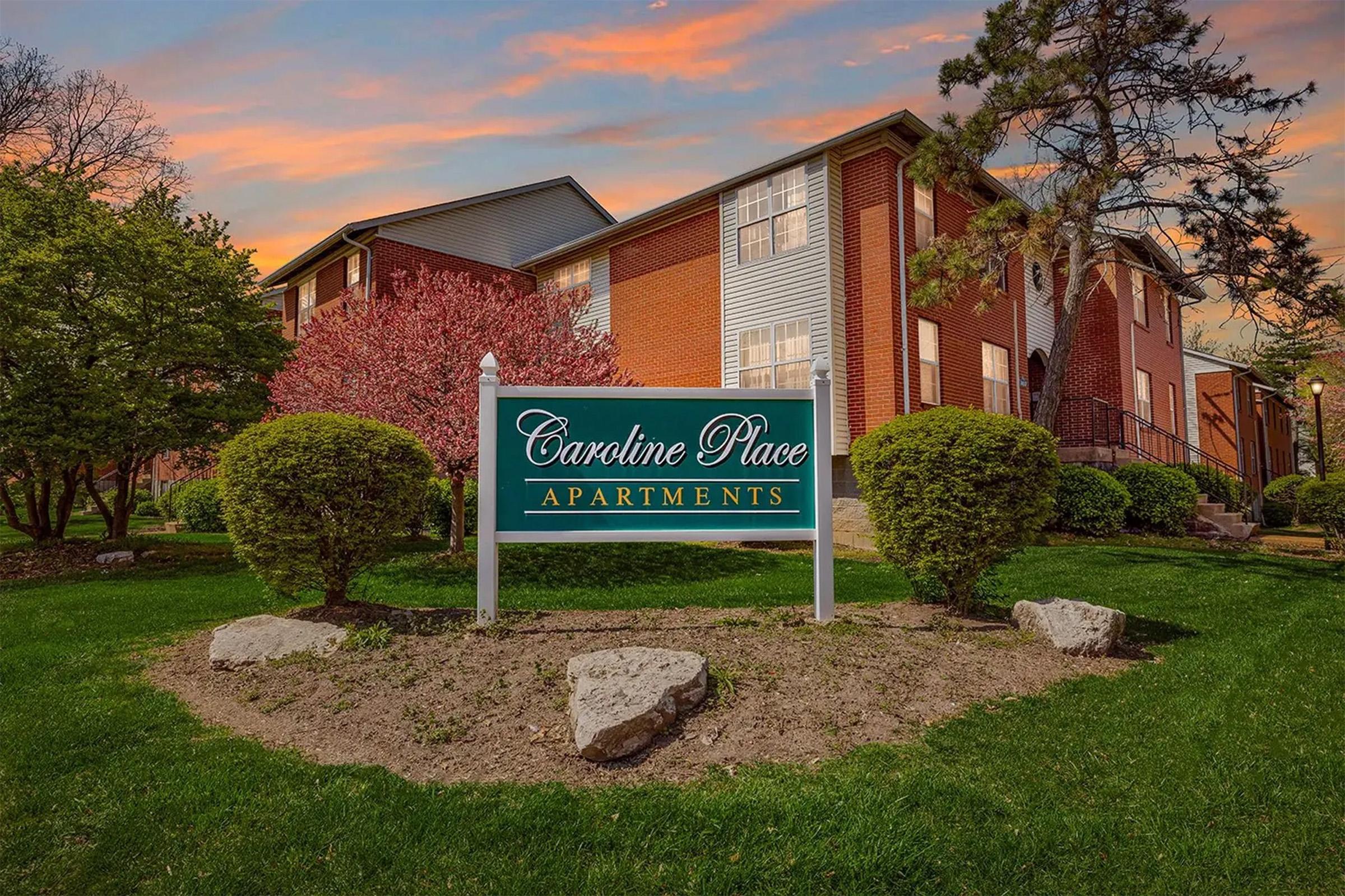 Sign for Caroline Place Apartments, surrounded by well-maintained landscaping, including bushes and small rocks. A sunset with pink and orange hues is visible in the background, adding a warm ambiance to the scene. The apartment building features a brick exterior and several windows.