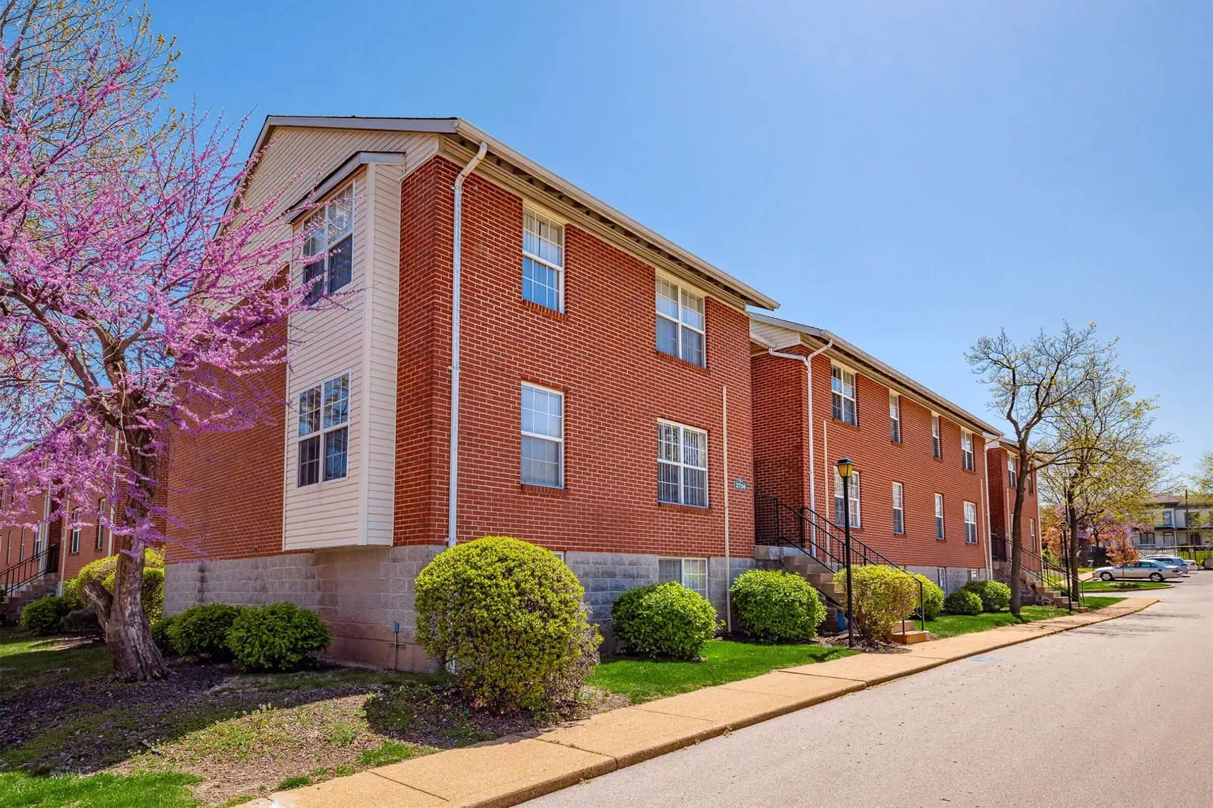 A brick apartment building with multiple windows, surrounded by well-maintained landscaping including a flowering tree and green shrubbery, under a clear blue sky. A sidewalk leads up to the building, emphasizing a tidy neighborhood setting.