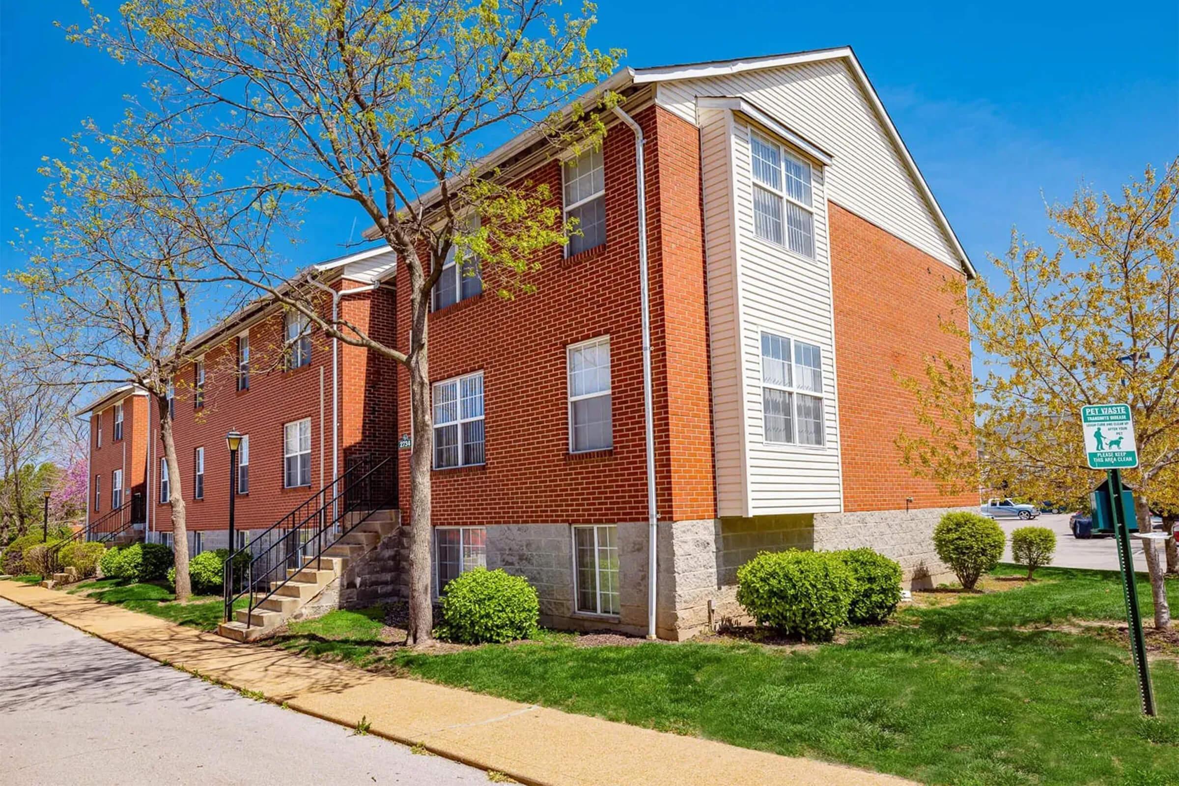 A brick apartment building with multiple windows and a staircase on the side, surrounded by green grass and small shrubs. Trees are budding, indicating early spring, under a clear blue sky. A sidewalk runs alongside the building, with a parking area visible in the background.