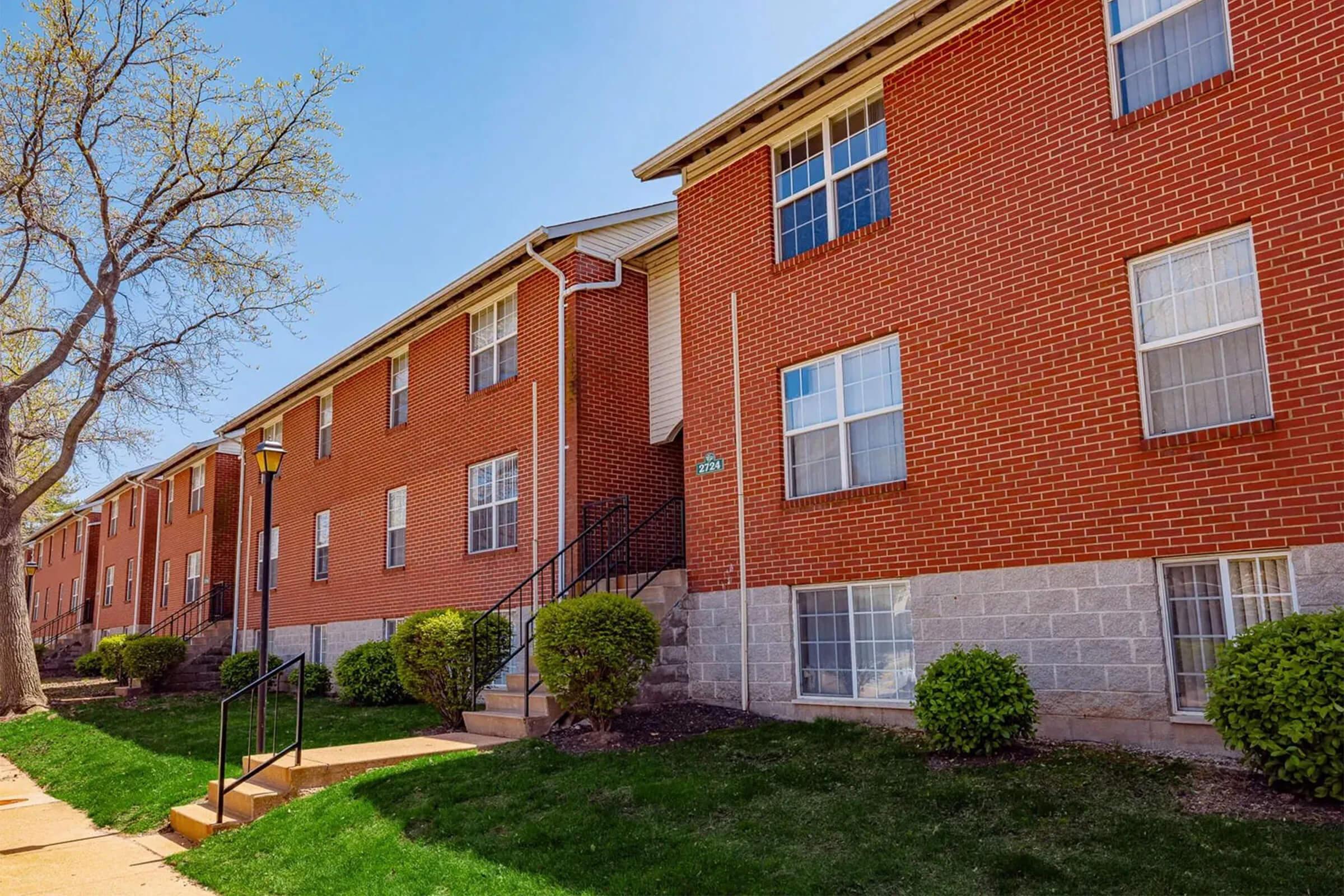 Brick apartment building with multiple windows, a set of stairs leading to the entrance, and neatly trimmed bushes in front. A tree is visible to the left, and the scene is set under a clear blue sky, indicating a sunny day.