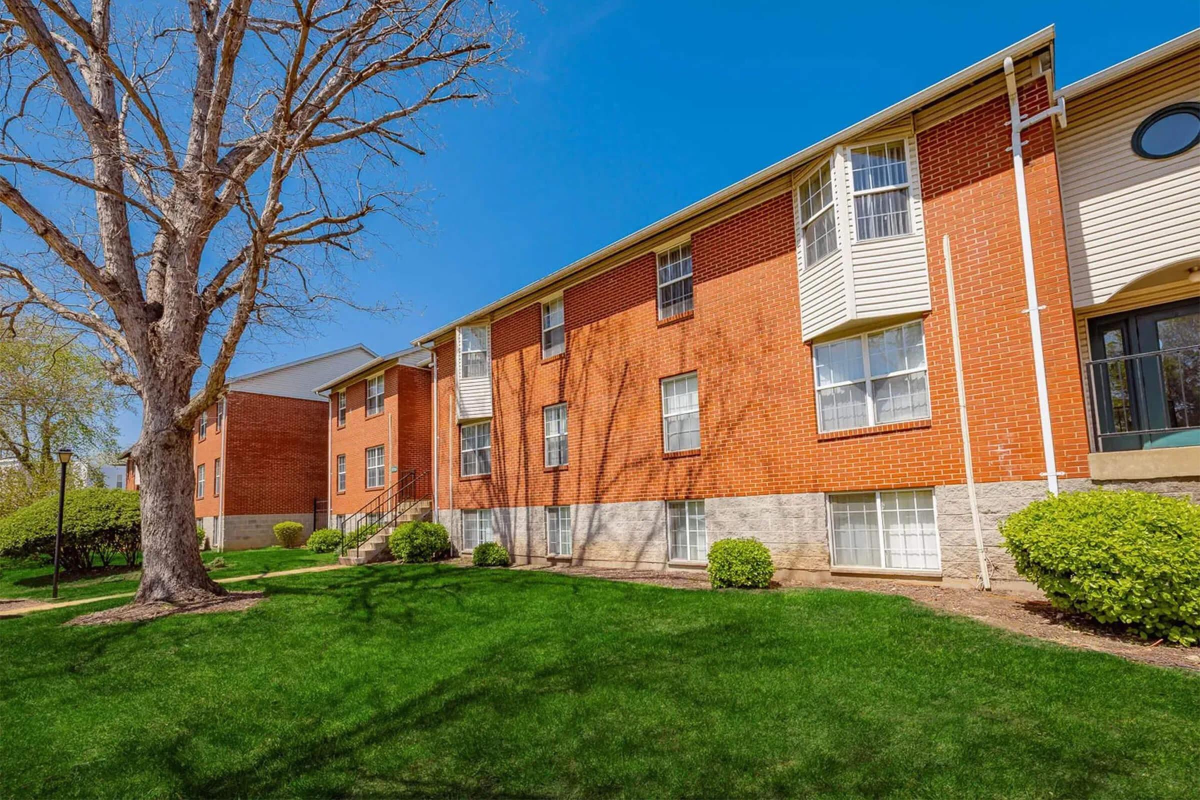 A red brick apartment building with multiple windows and a grassy area in front. A large tree is present on the left side, and there are small bushes along the building's foundation. The sky is clear and blue, suggesting a sunny day.