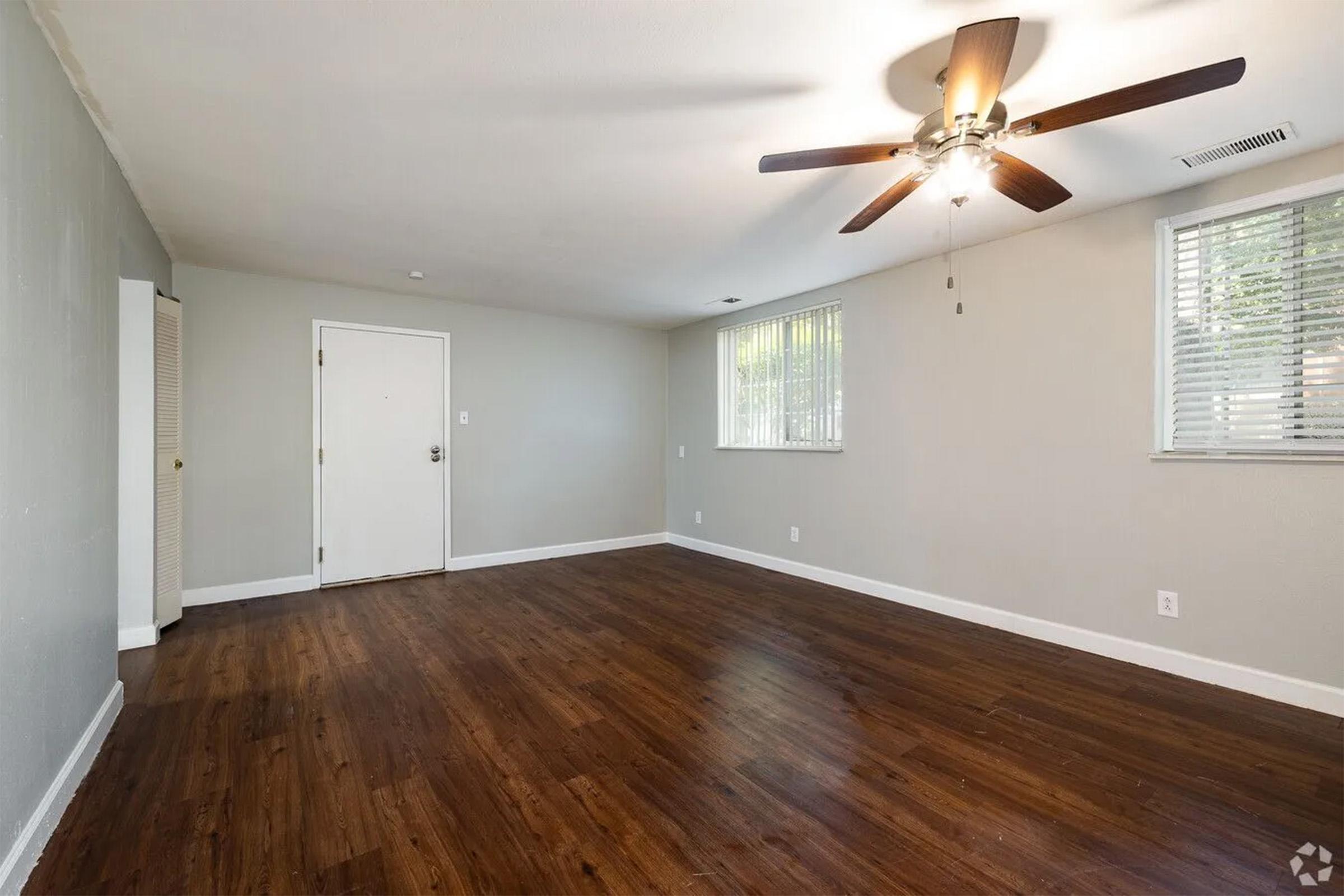 A spacious, empty living room with light gray walls and a hardwood floor. There is a ceiling fan with wooden blades, a closed door on one wall, and two windows with blinds letting in natural light. The room is well-lit and clean, creating an inviting atmosphere.