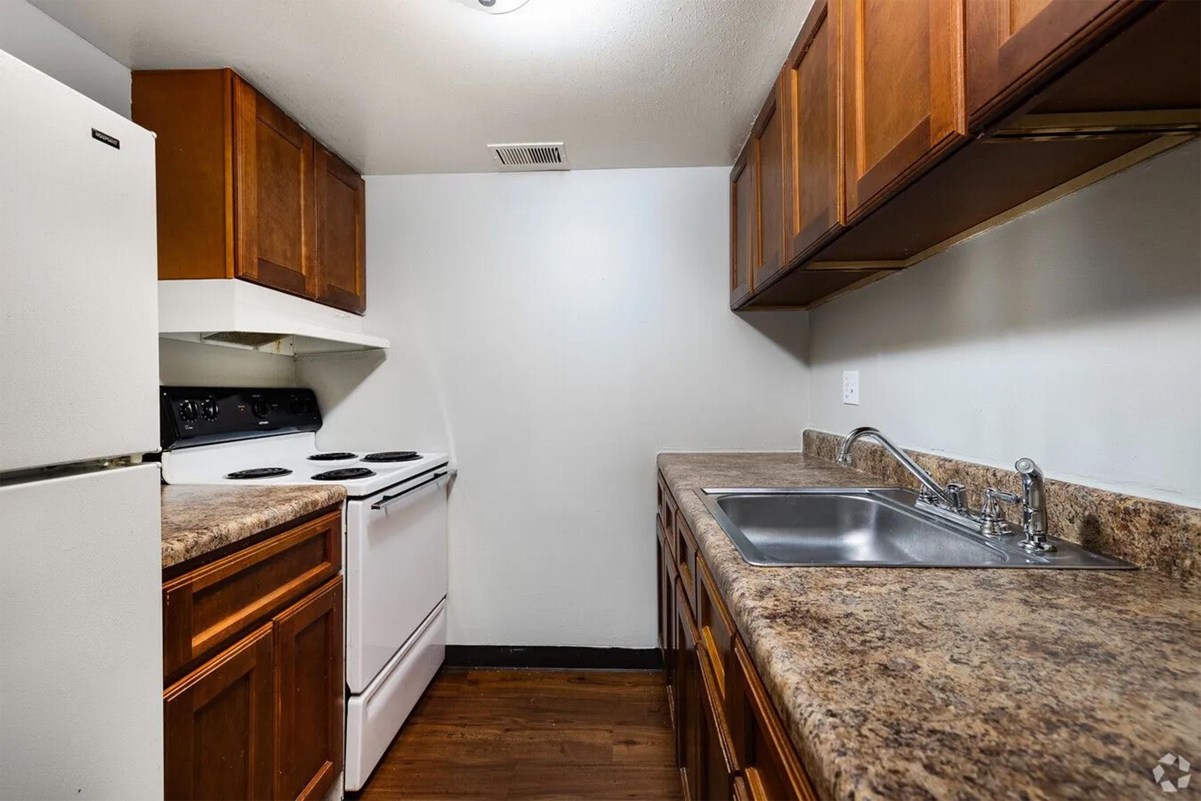 A small kitchen featuring wooden cabinets, a white refrigerator, a black stove with an oven, and a stainless steel sink. The countertops are a beige stone texture, and the walls are light-colored, creating a clean and simple look.