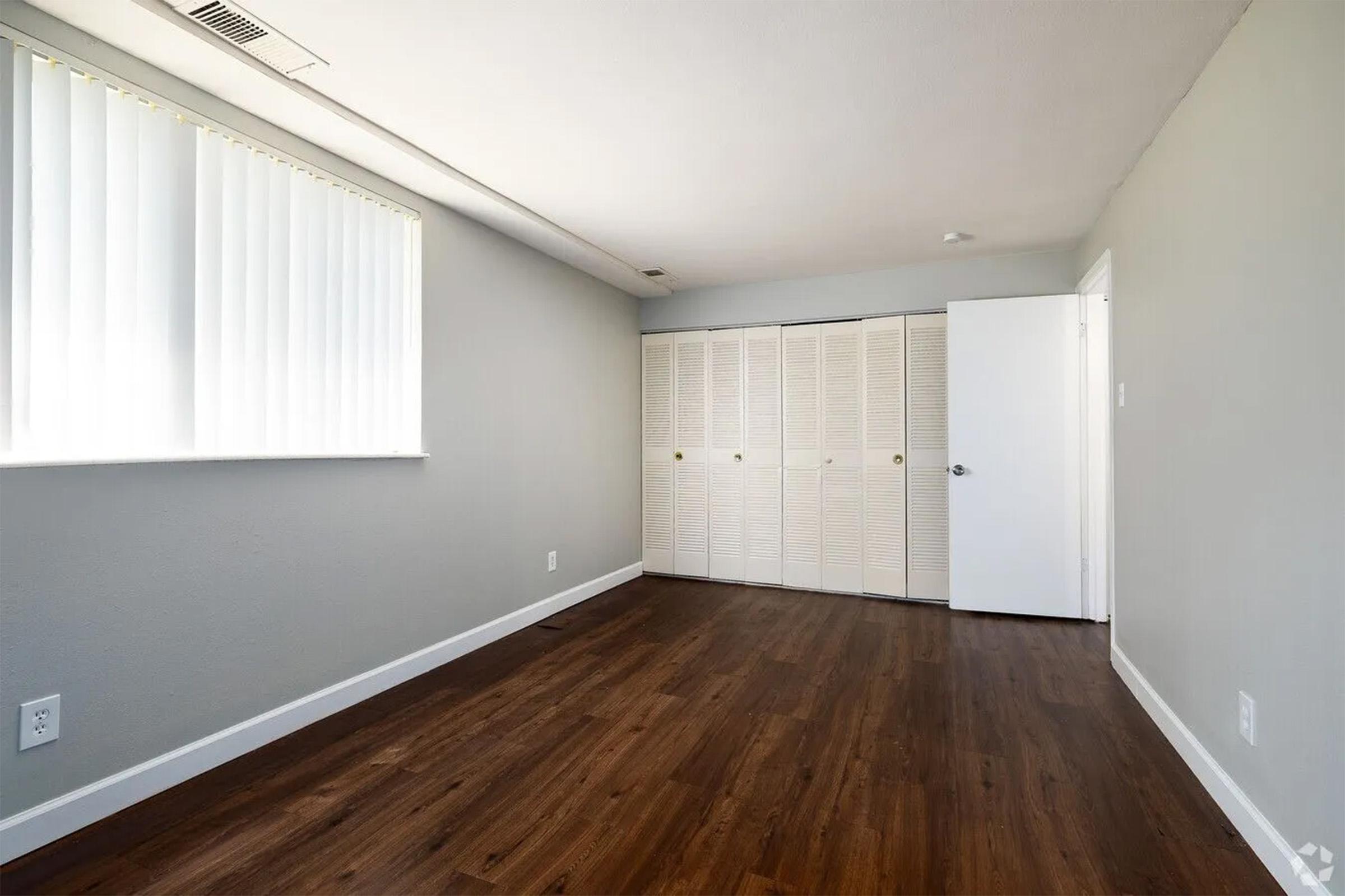 A minimally furnished room with light gray walls and a wooden floor. There is a long window covered with blinds on one side, and a closet with multiple doors against the opposite wall. The room is well-lit, creating a simple and open atmosphere.