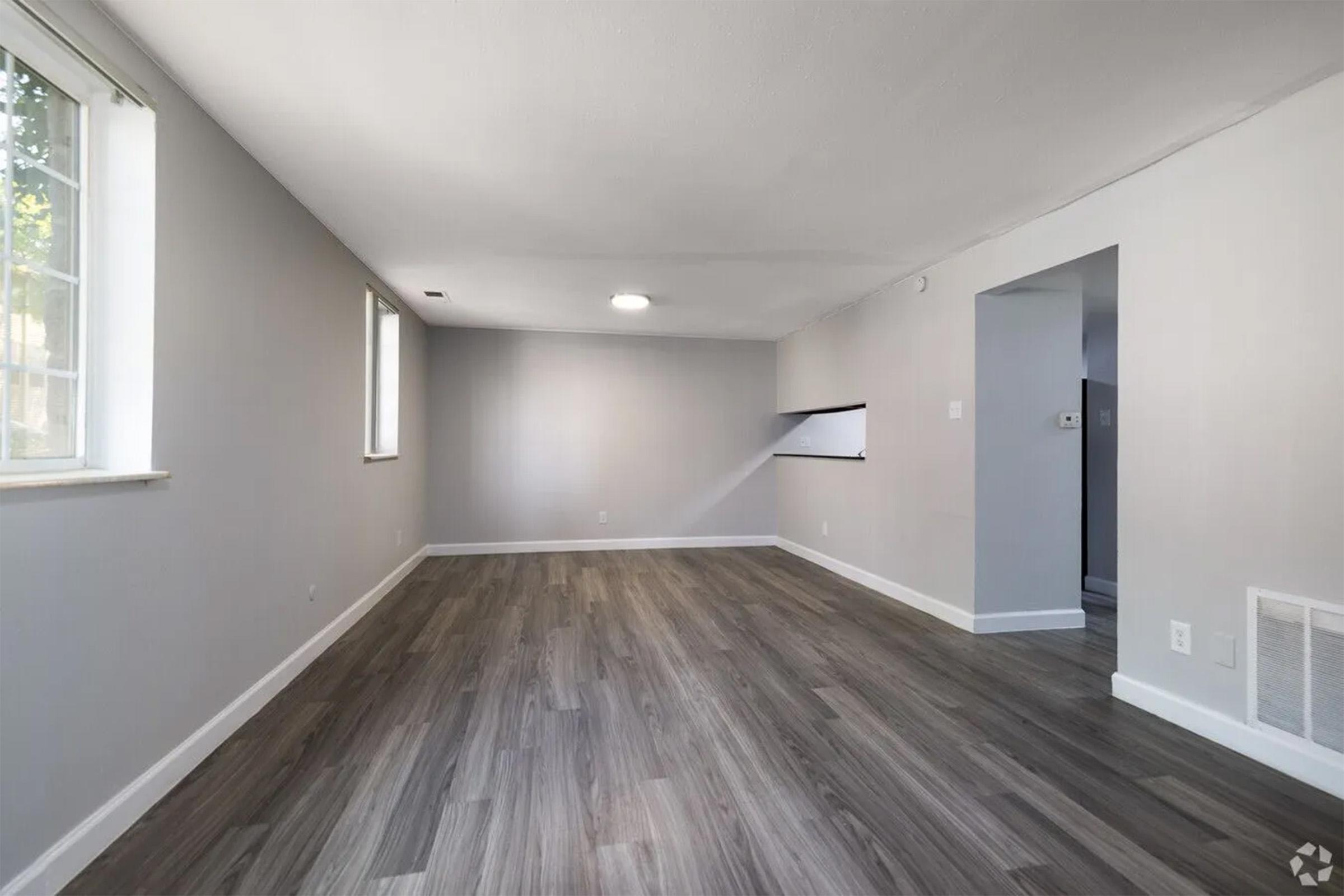 A bright, empty room with gray walls and laminate flooring. A large window allows natural light to enter, creating an open and airy feel. The layout features a wall cutout leading to another space, with no furniture, emphasizing the room's spaciousness.