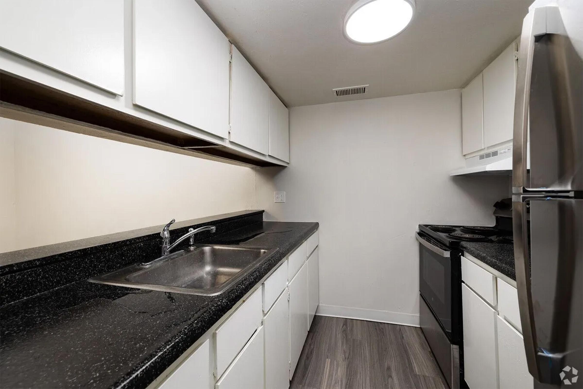 A modern kitchen featuring white cabinetry, a dark granite countertop, a stainless steel sink, and a black stove and refrigerator. The space is well-lit with a ceiling light and has vinyl flooring, creating a clean and functional cooking area.