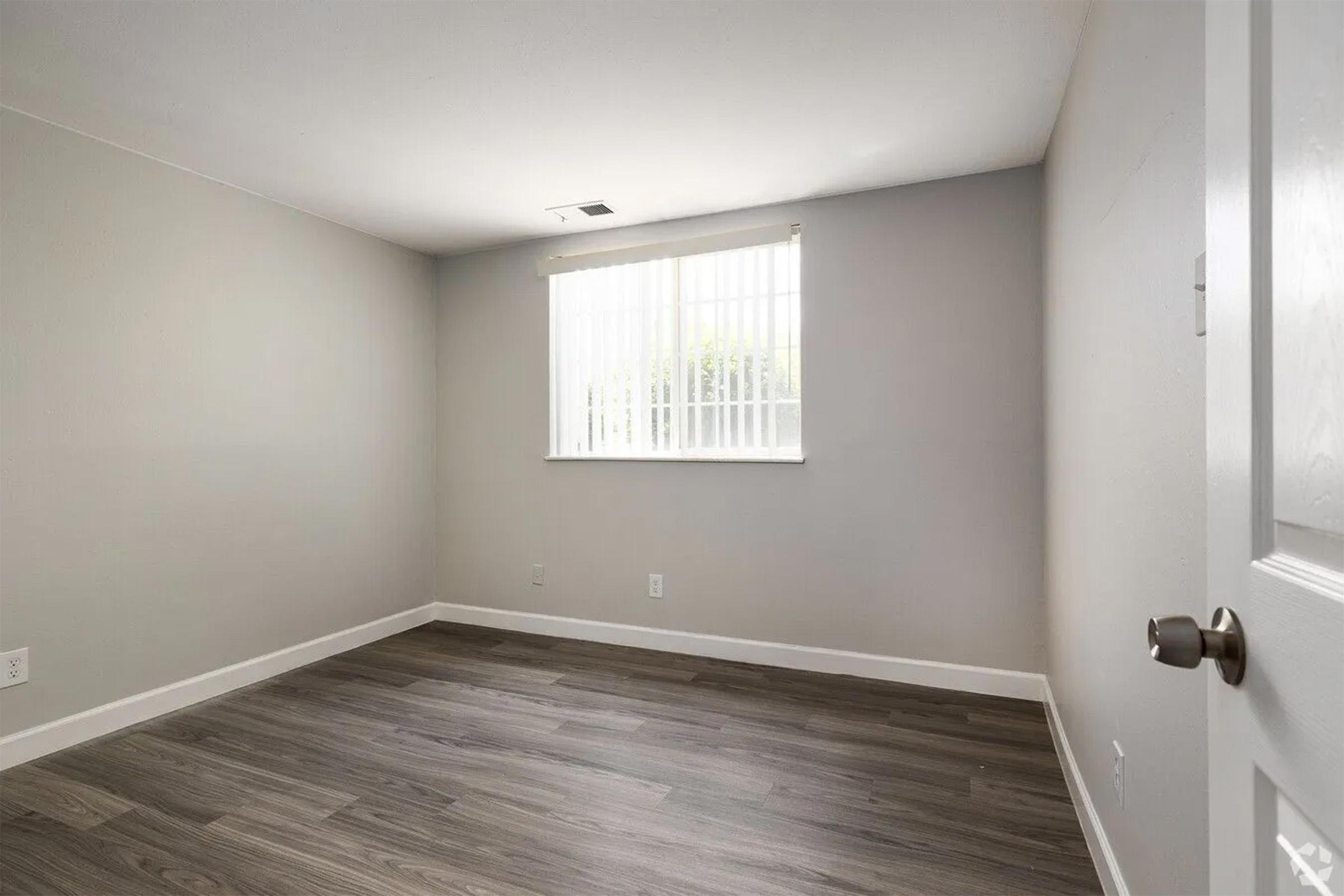 Empty room with light gray walls and a wooden floor. A single window with blinds lets in natural light. The space is uncluttered, featuring a white door on the right side. The room is well-lit and appears to be freshly painted.