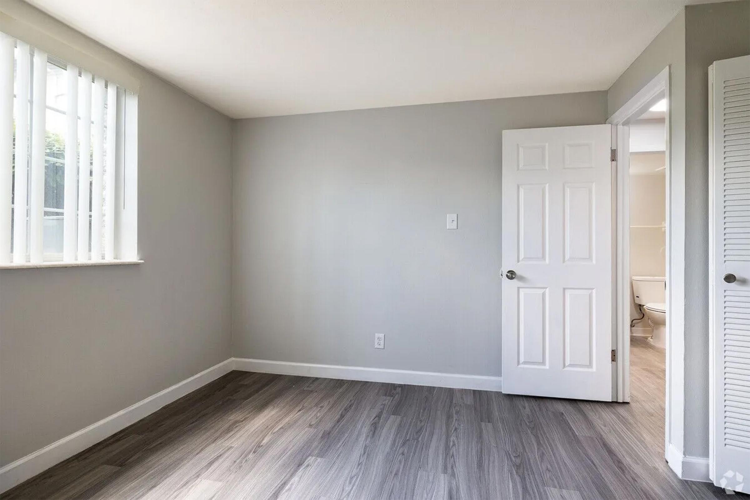 A small, empty room with light gray walls and laminate flooring. It features a window with vertical blinds on the left side. A doorway leads to another room on the right, partially visible. The overall space is bright and minimalistic, offering a clean and open feel.
