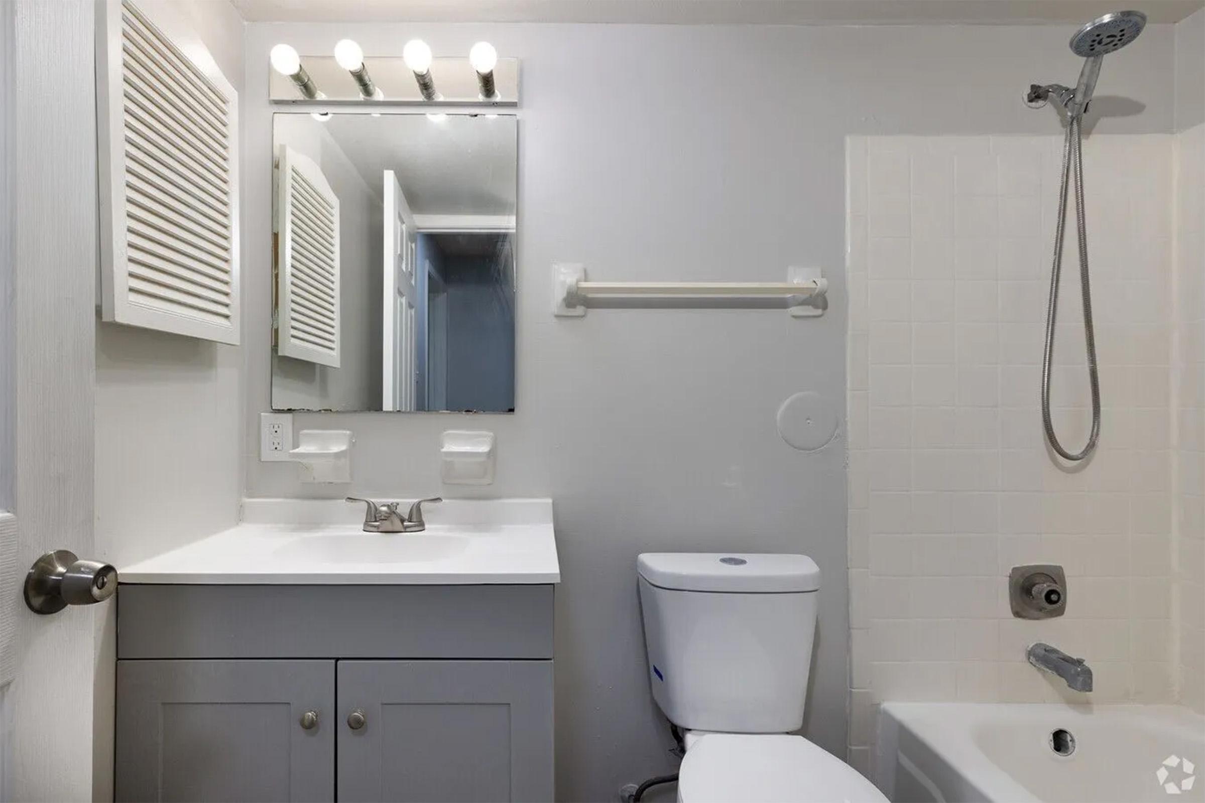 A clean, modern bathroom featuring a white bathtub with a showerhead, a toilet, and a vanity with a sink. There is a large mirror above the sink, and four light fixtures are mounted above it. A towel rack is attached to the wall, and the room has neutral-colored walls and tiled flooring.