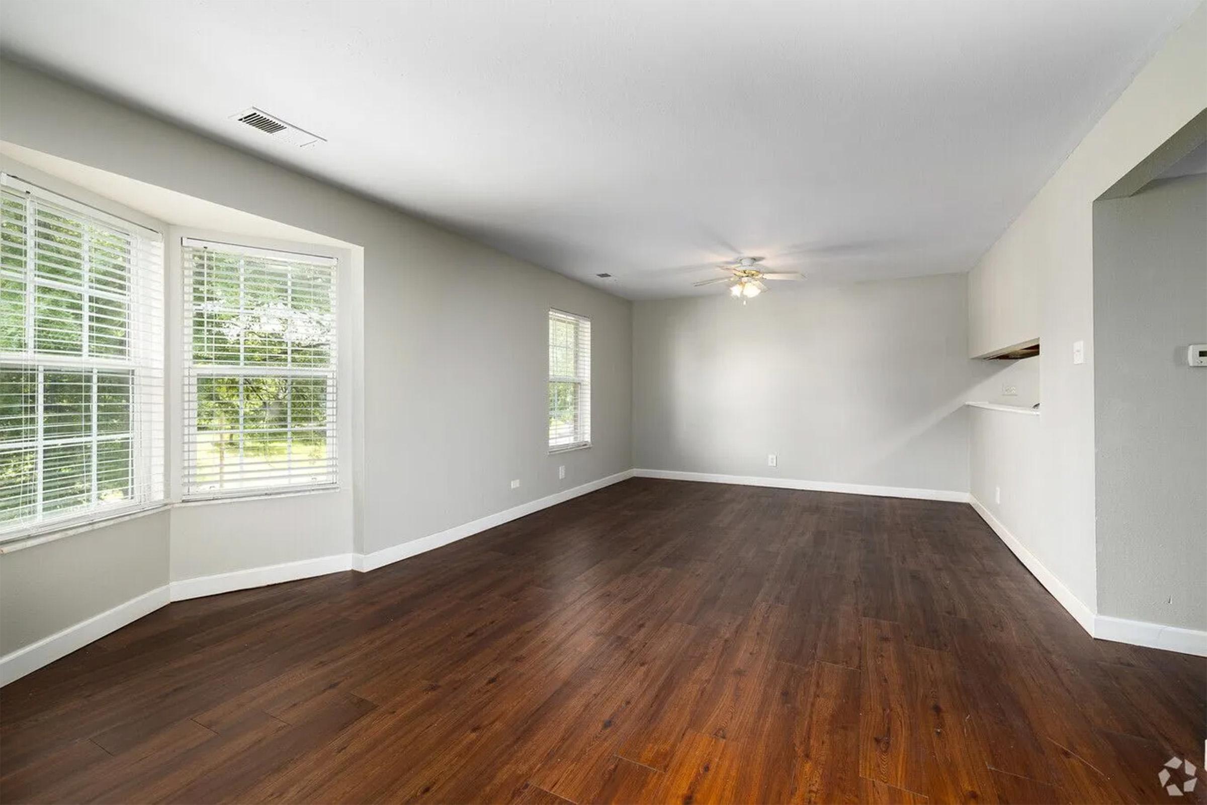 A spacious, empty room with hardwood flooring, large windows letting in natural light, and neutral-colored walls. The ceiling features a light fixture, and there is an open area leading to a kitchen space. The atmosphere is bright and airy, suitable for various uses.
