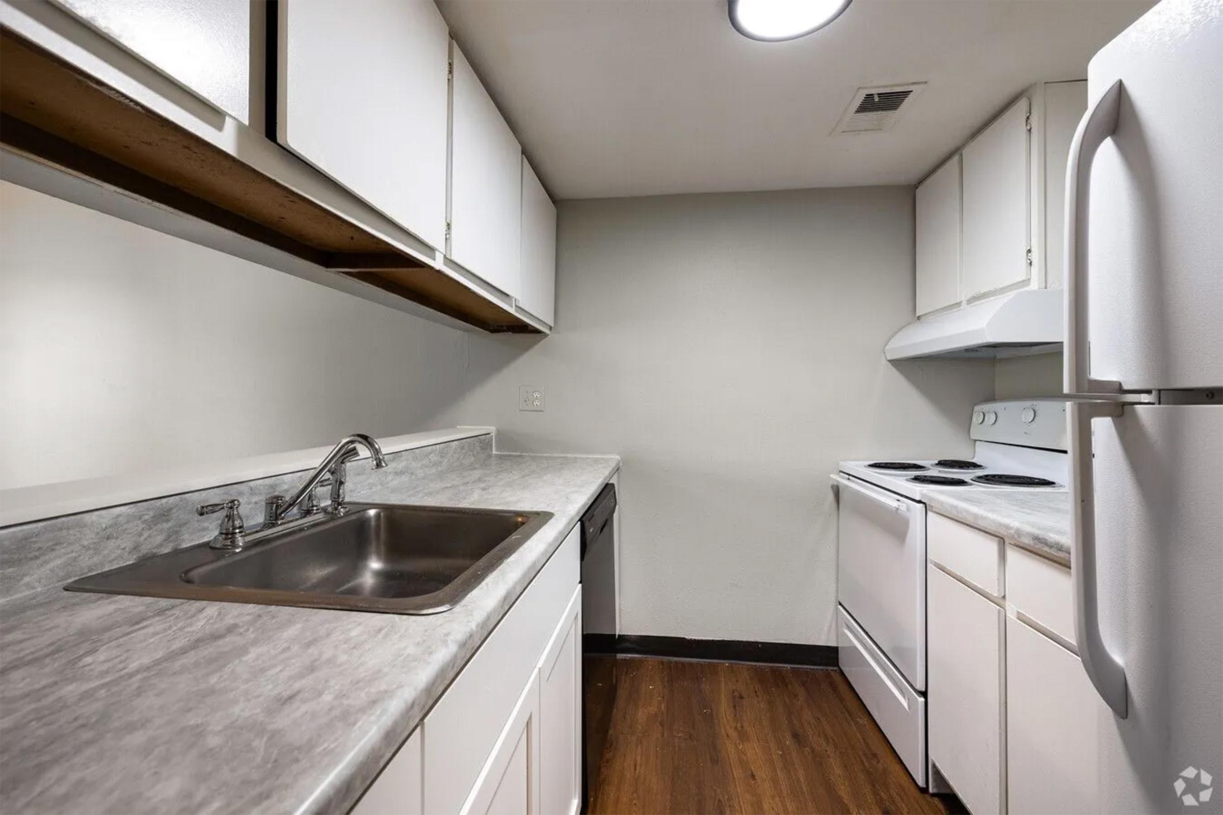 A modern kitchen featuring white cabinetry, a stainless steel sink, and a countertop with a marble-like finish. The kitchen is equipped with a stove, an oven, and a refrigerator. The flooring is dark wood, and the walls are painted a light color, creating a bright and spacious atmosphere.