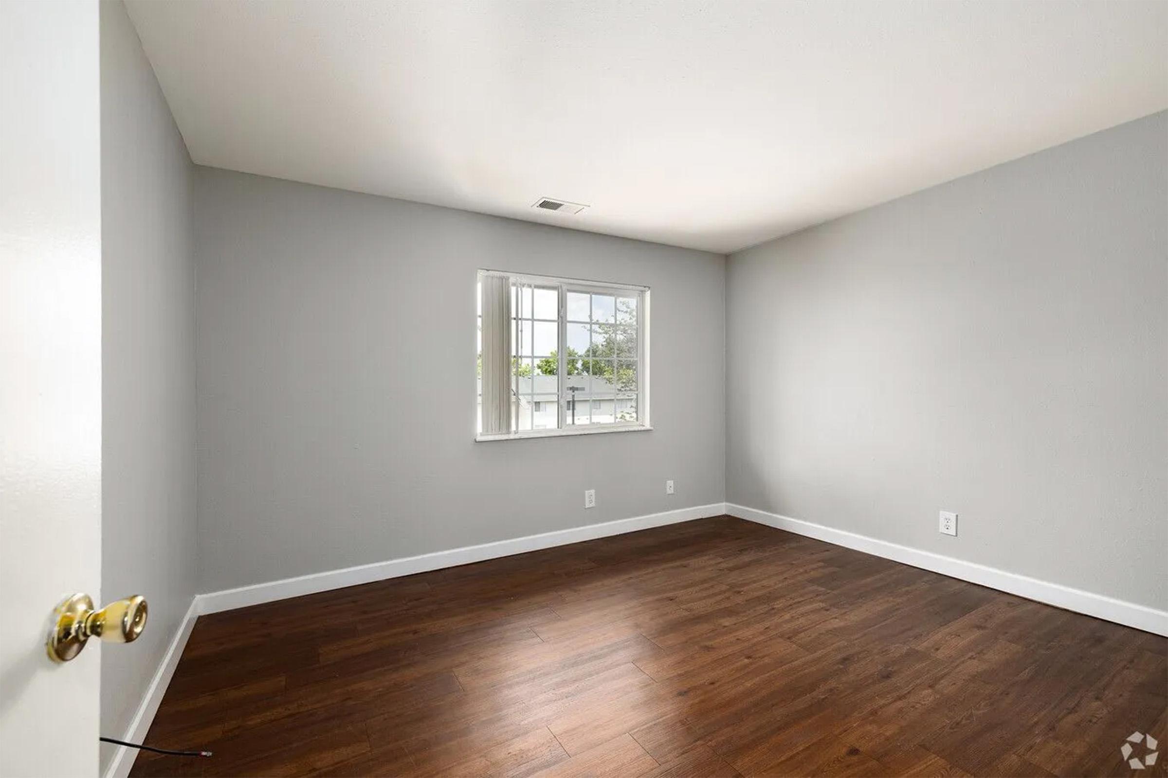 Empty room with gray walls and a window letting in natural light. The floor is made of hardwood, and there's a simple door with a round handle. The space appears clean and uncluttered, suitable for various uses.