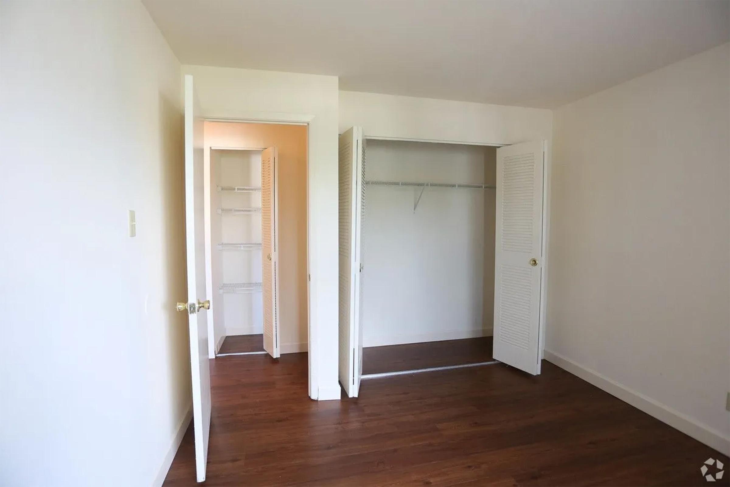A spacious, empty room with light-colored walls and hardwood flooring. There are two open doors leading to a small closet with a hanging rod and a second door leading to another room or hallway. The room is well-lit, creating a clean and airy atmosphere.