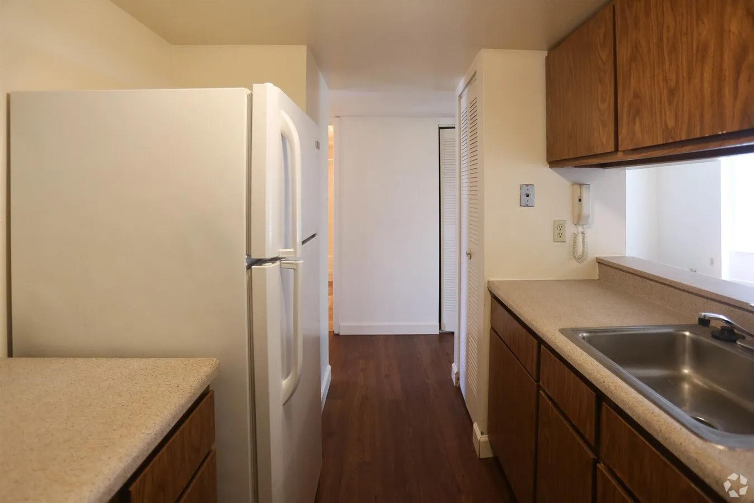 A simple kitchen with wooden cabinets, a white refrigerator, and a countertop. A sink is visible next to the window. The room has wooden flooring and leads to a hallway with a closed door in the background. The overall aesthetic is bright and functional.