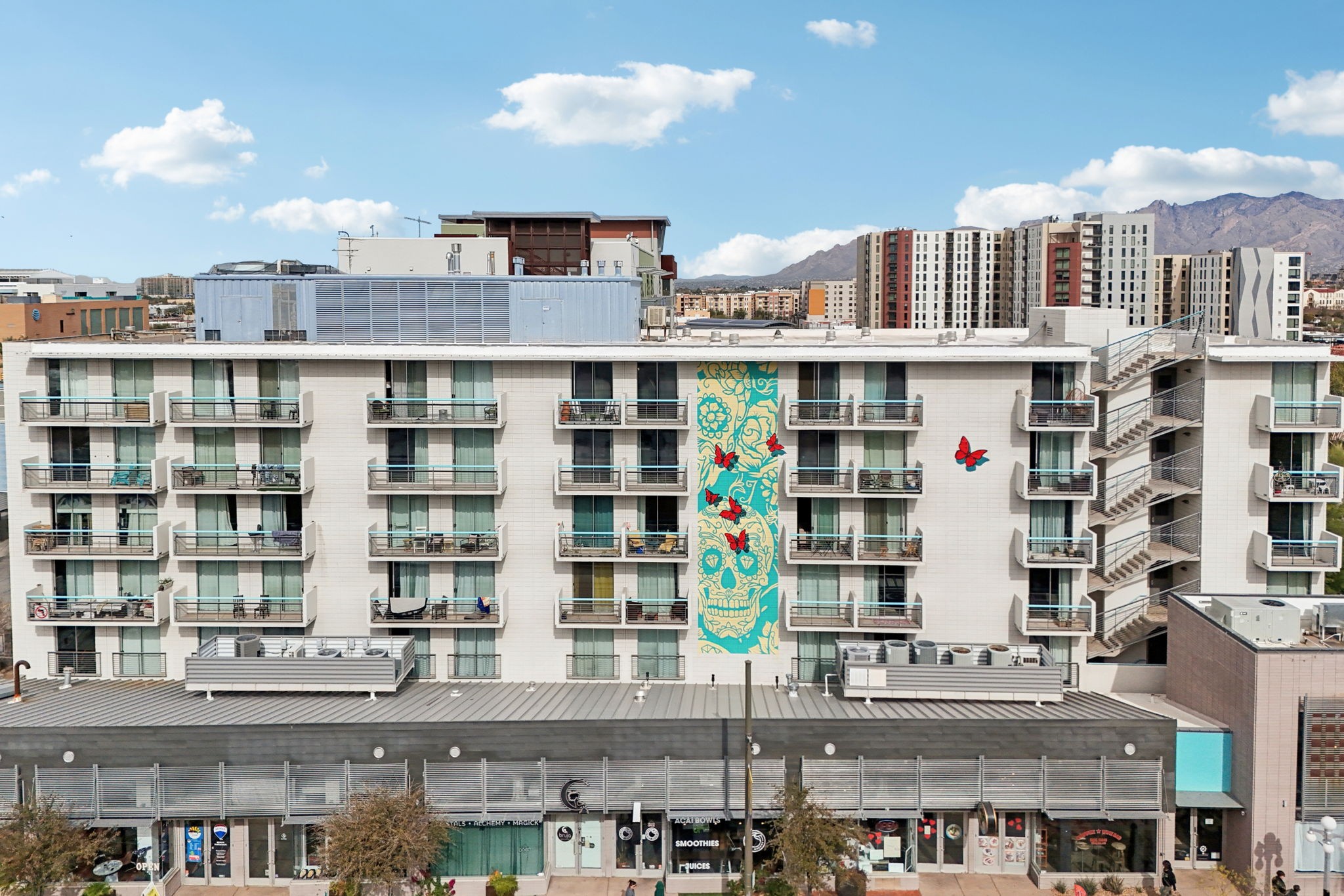 A multi-story building with balconies, featuring a large colorful mural of a sugar skull surrounded by butterflies on one side. The scene is set against a clear blue sky with a few clouds, showcasing a modern urban environment. Nearby buildings and mountains are visible in the background.