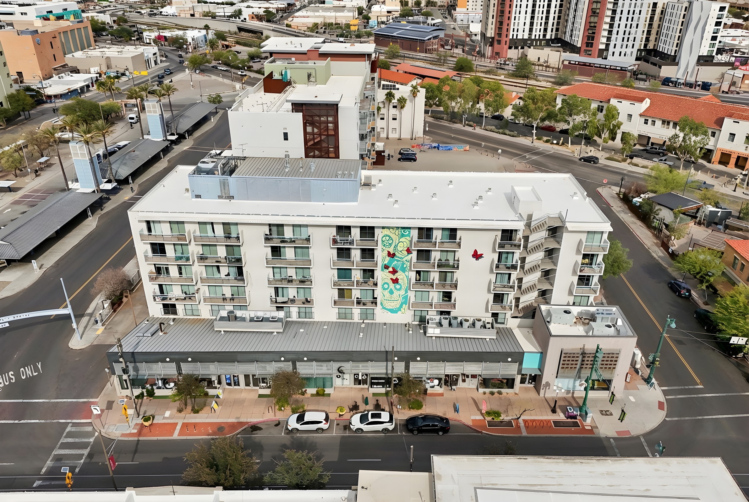 Aerial view of a modern multi-story building with a colorful mural on the side. The ground level features storefronts and parked cars along a city street. Surrounding area includes other buildings and greenery, with a clear sky in the background.