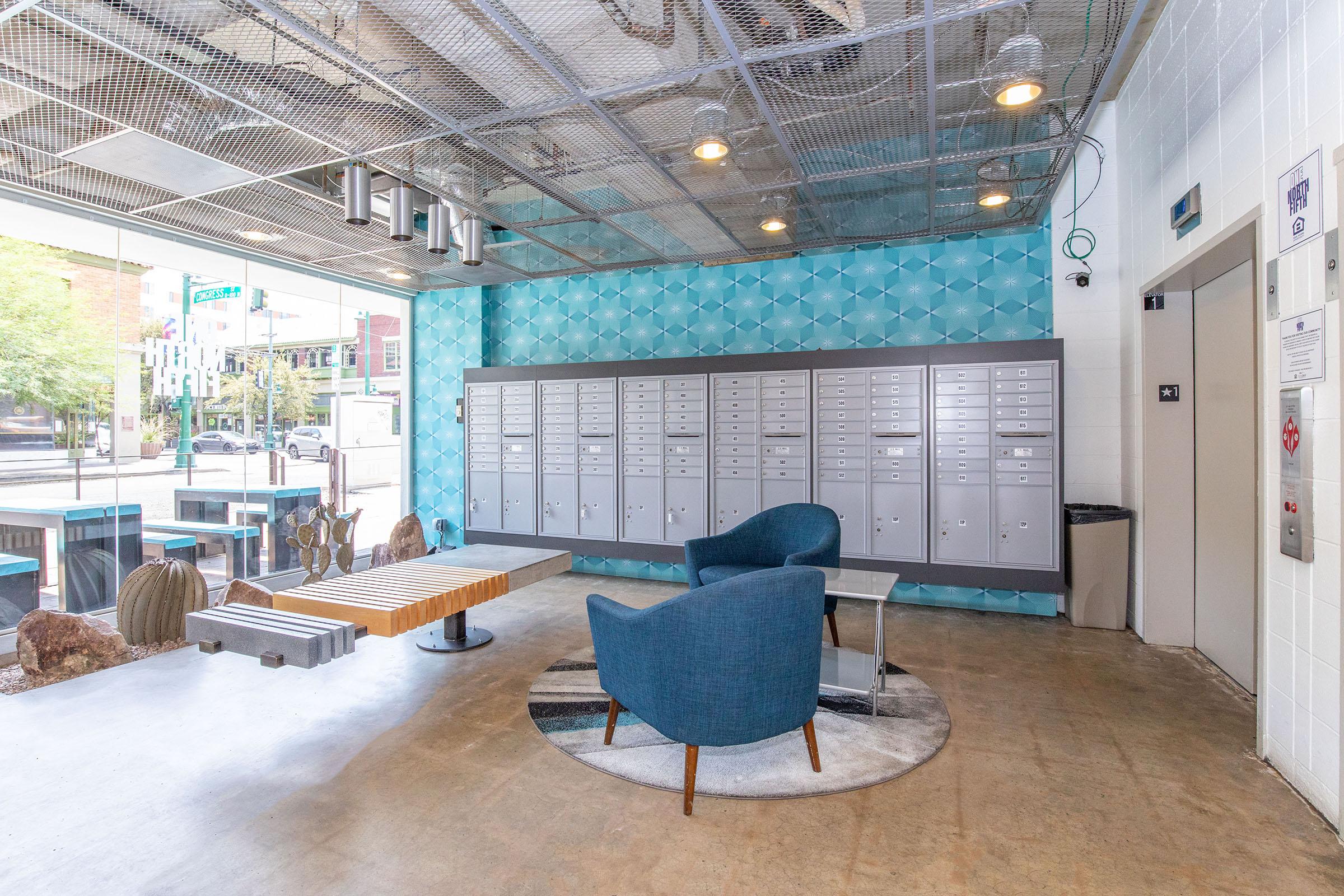 A modern lobby area featuring blue accent walls, two comfortable blue chairs, a round coffee table, and a large circular rug. In the background, there are multiple mailboxes and an elevator door. The space has a bright and welcoming atmosphere, with large windows allowing natural light to enter.